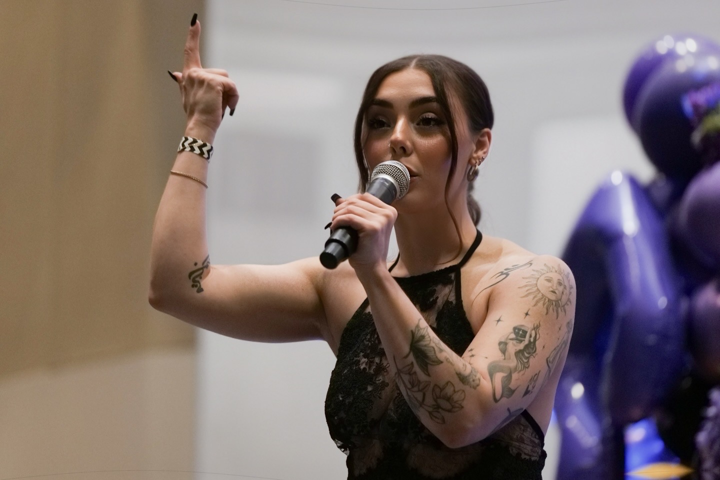 Woman with tattoos speaking into a microphone, gesturing with her right hand, wearing a black lace top, in front of balloons.