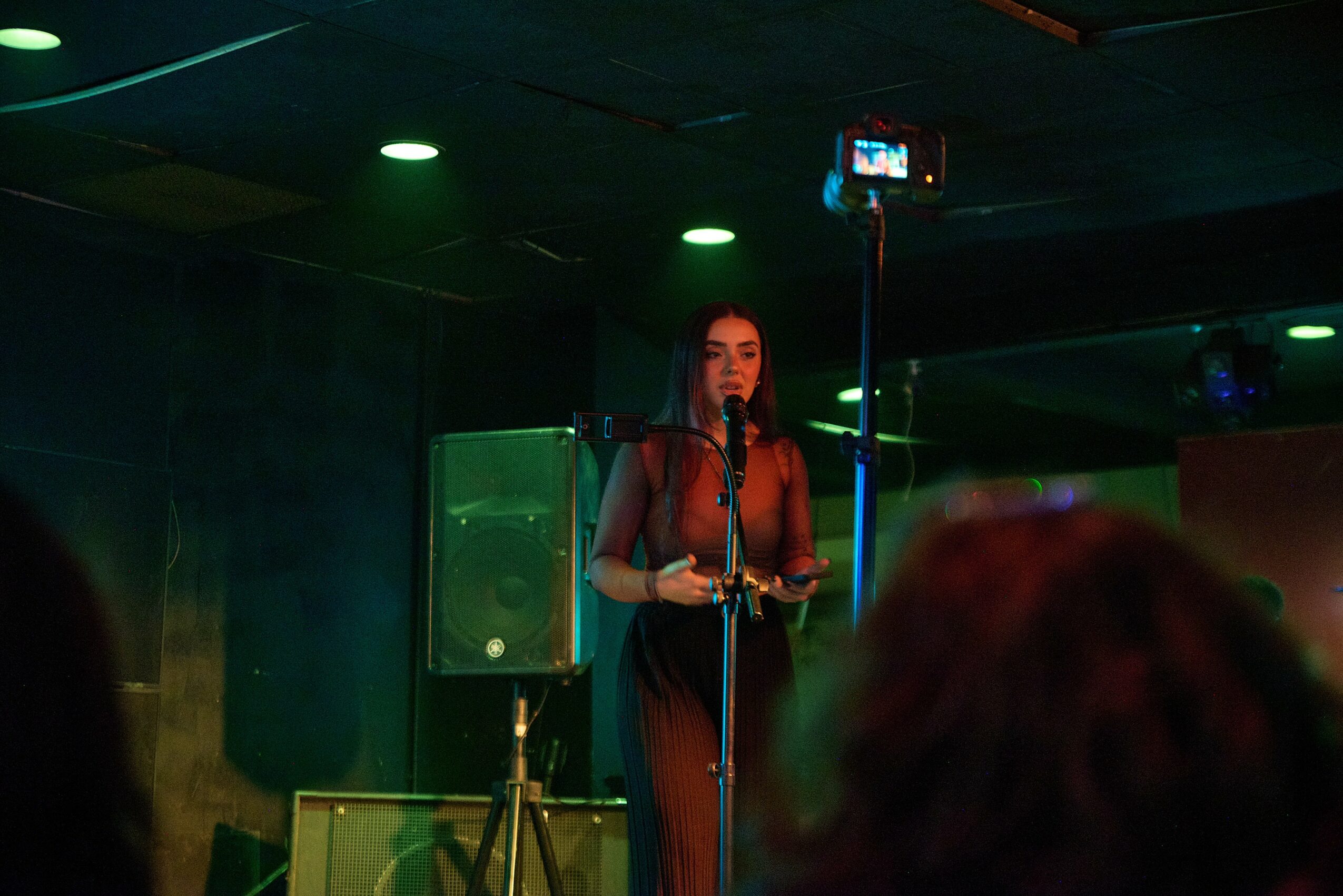 A woman stands on stage with a microphone, speaking or singing, in a dimly lit venue with speakers and a camera overhead.