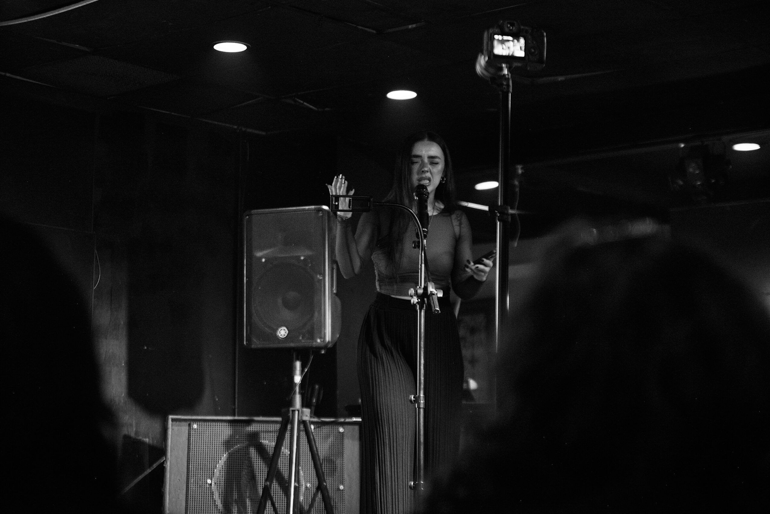 A woman stands on stage, speaking or singing into a microphone, with a speaker behind her and a dark background.