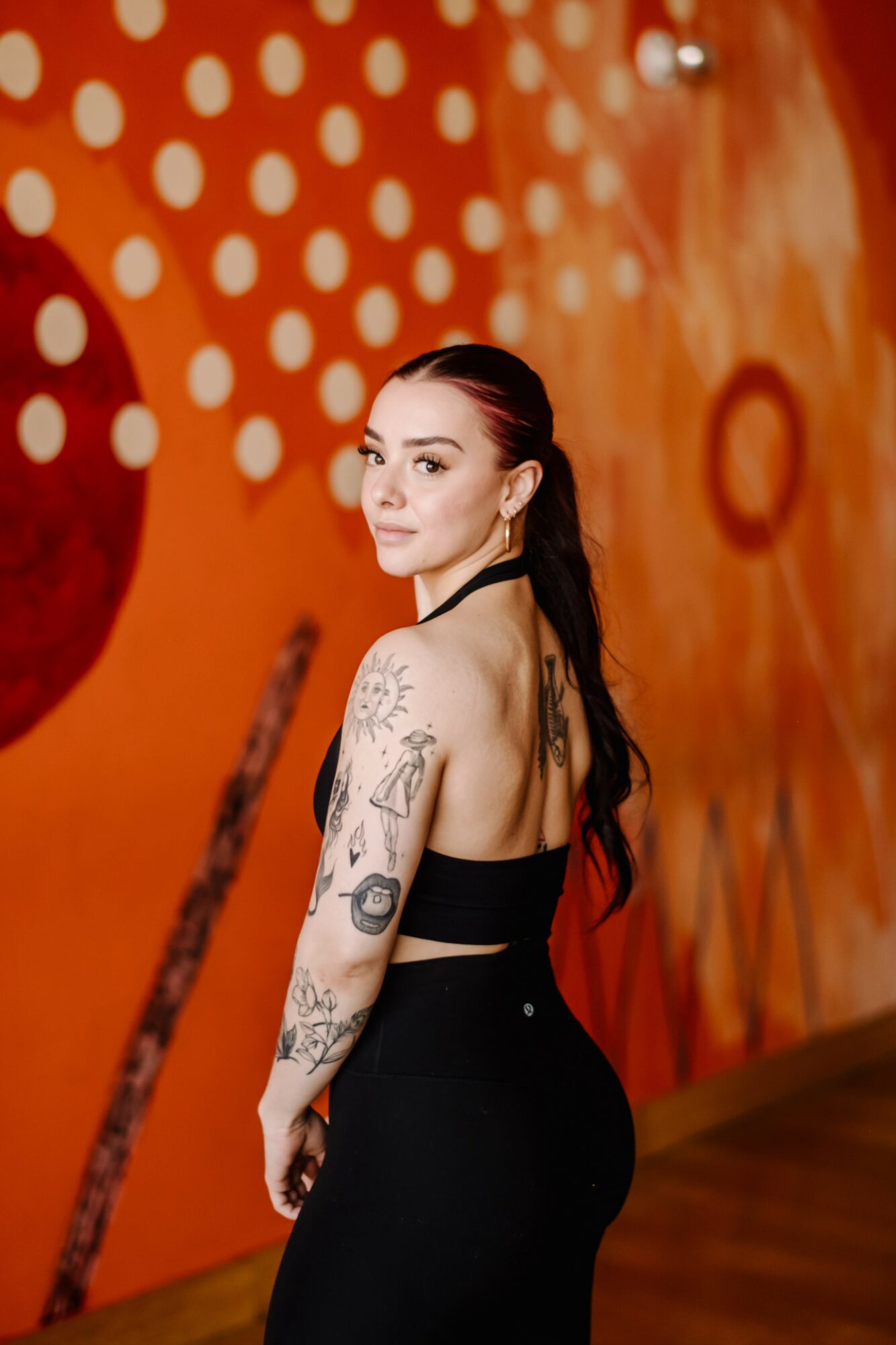Young woman with long dark hair and tattoos on her arm, standing against an orange wall with white dots.