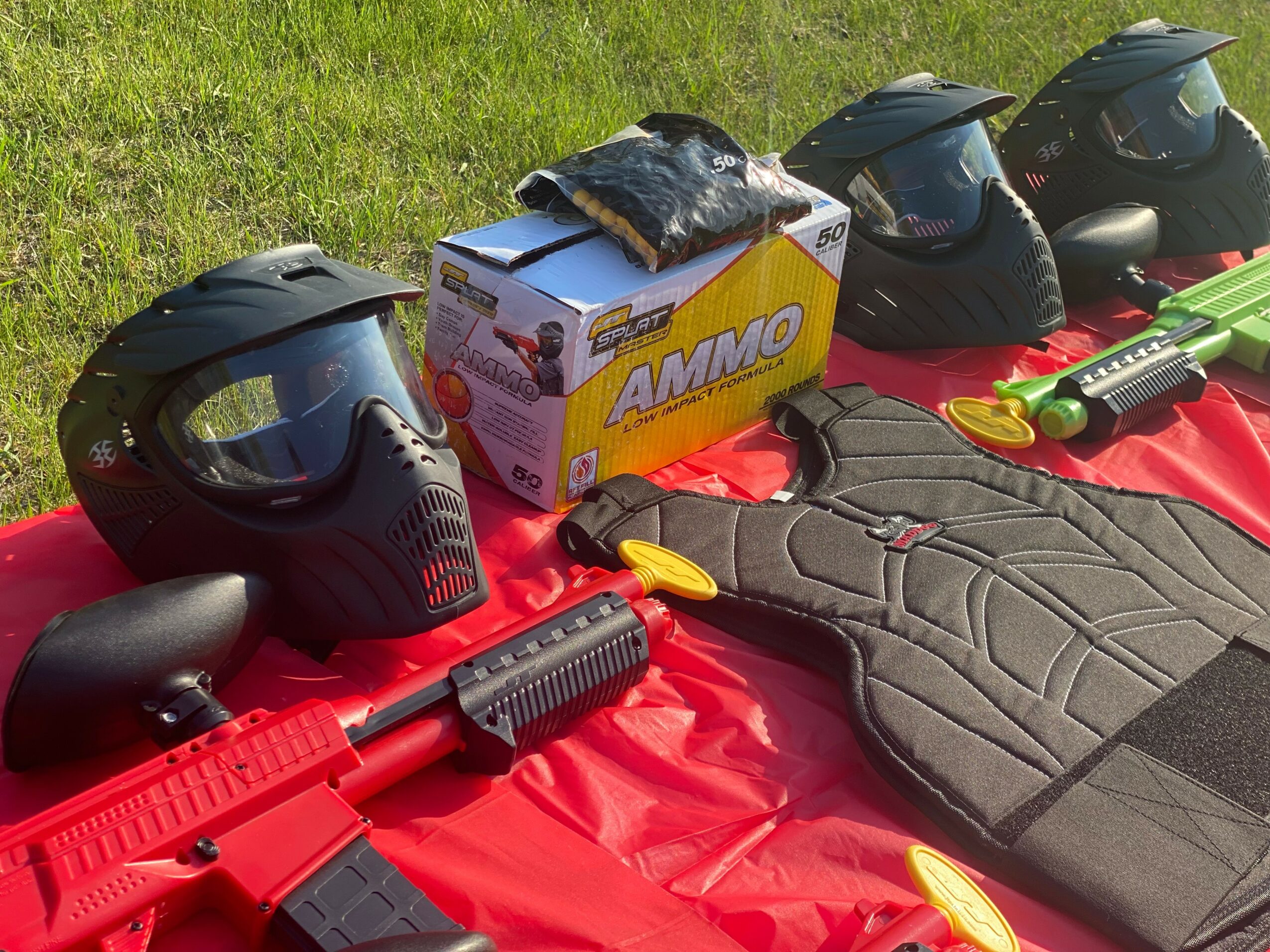 Four paintball masks, a box labeled 'AMMO,' and paintball guns on a red surface outdoors.