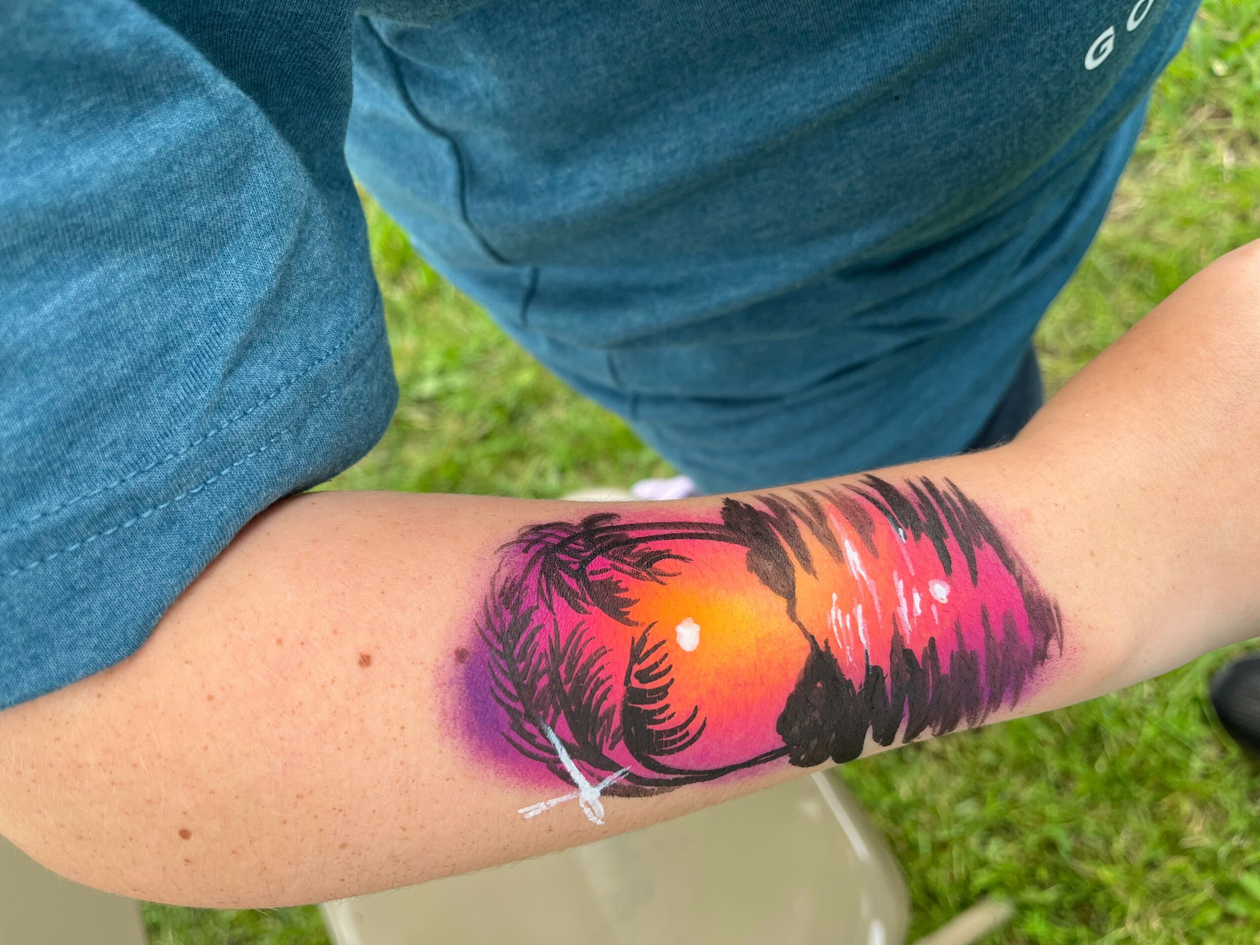 Arm with colorful sunset tattoo featuring palm trees and ocean waves, outdoors on grass, person wearing blue shirt