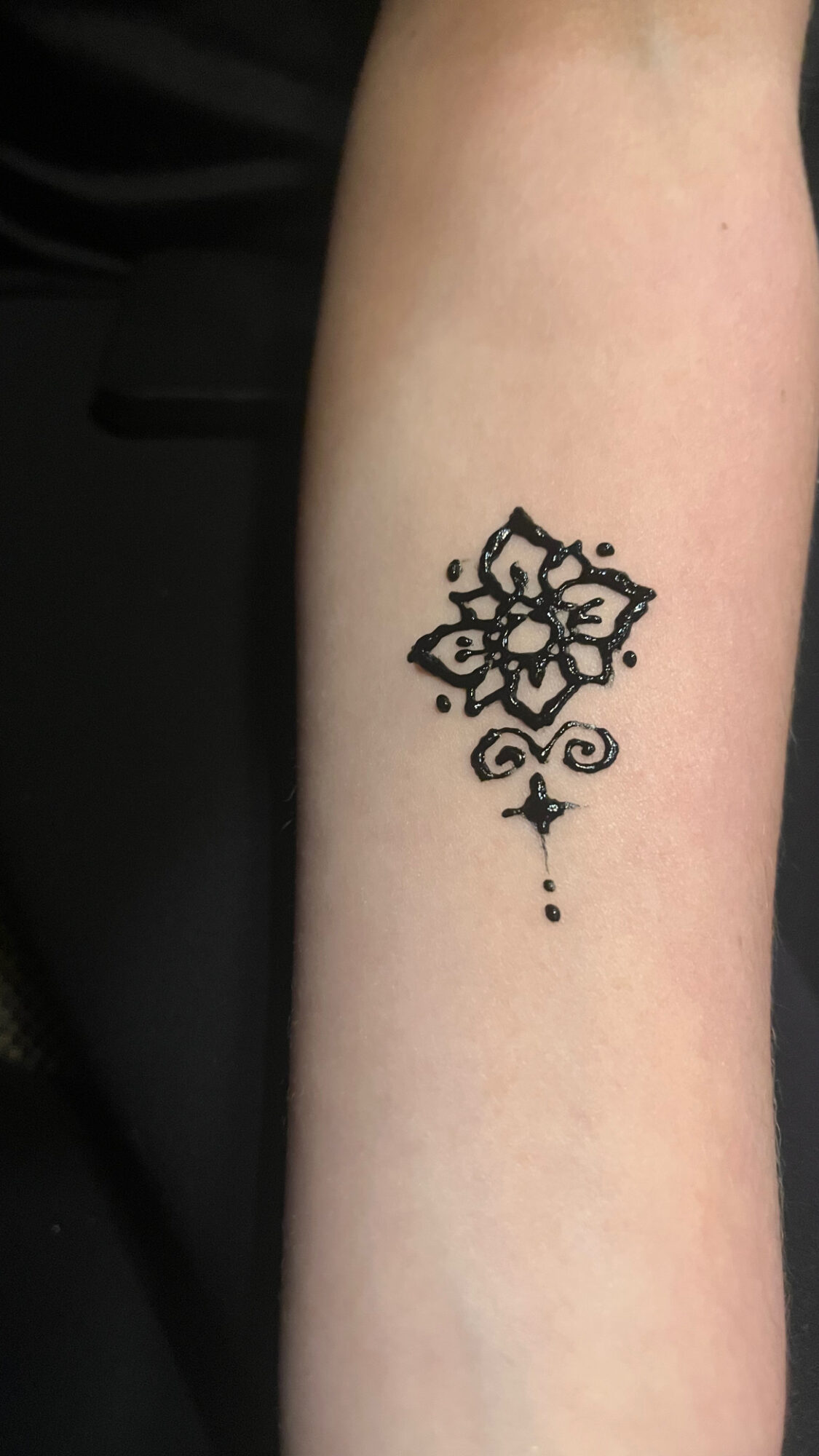 Tattoo of a symmetrical geometric design with a small cross and dots below on a person's forearm.