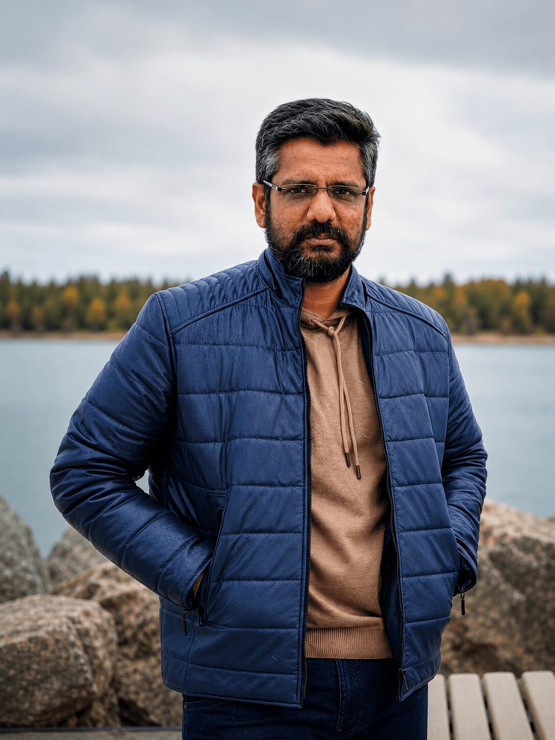 Man with glasses and beard standing outdoors near water, wearing a blue jacket and brown hoodie, with trees in background.