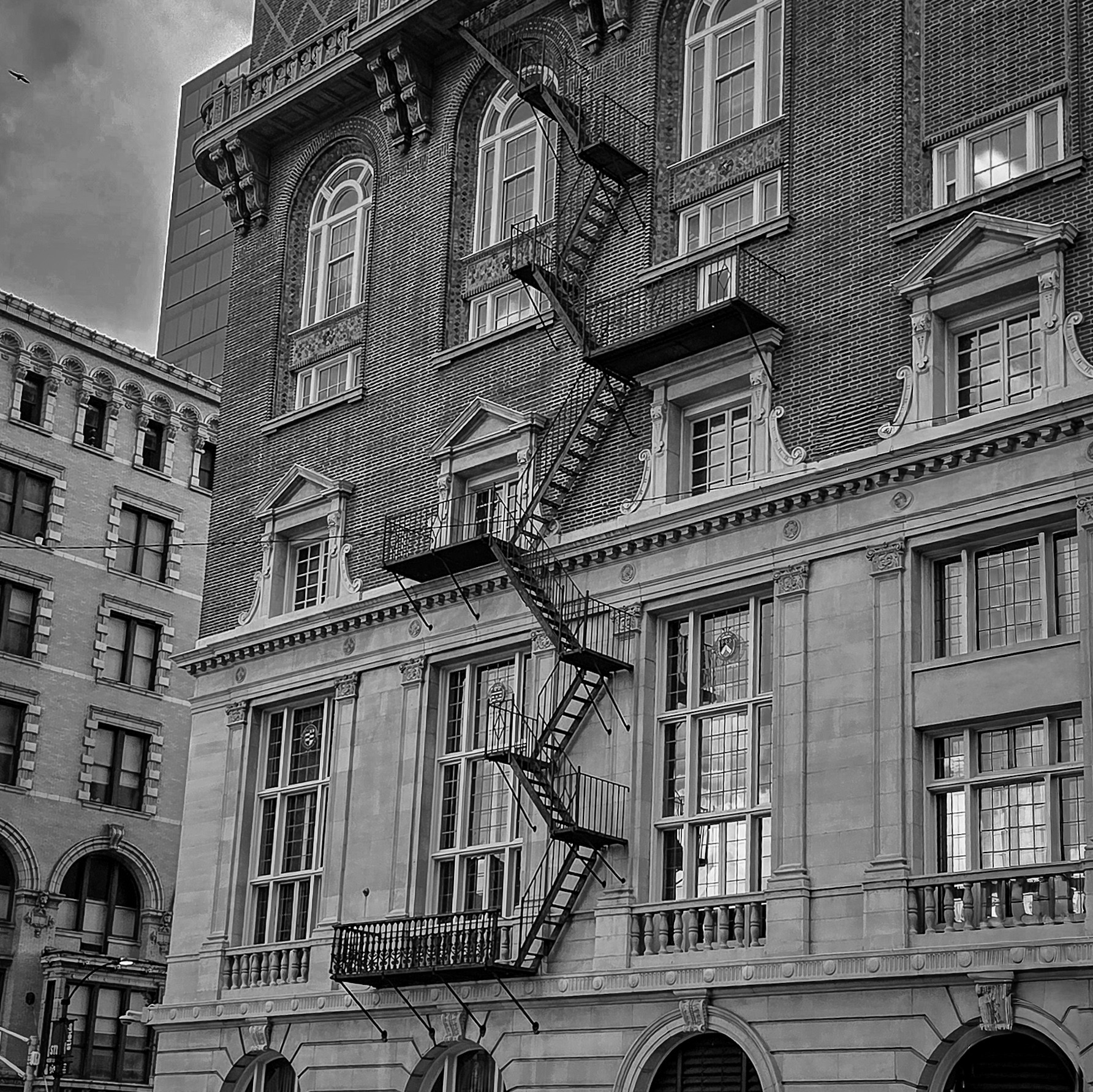 Multi-story brick building with fire escape zigzagging down the facade, windows with decorative frames, and a balcony, in black and white.