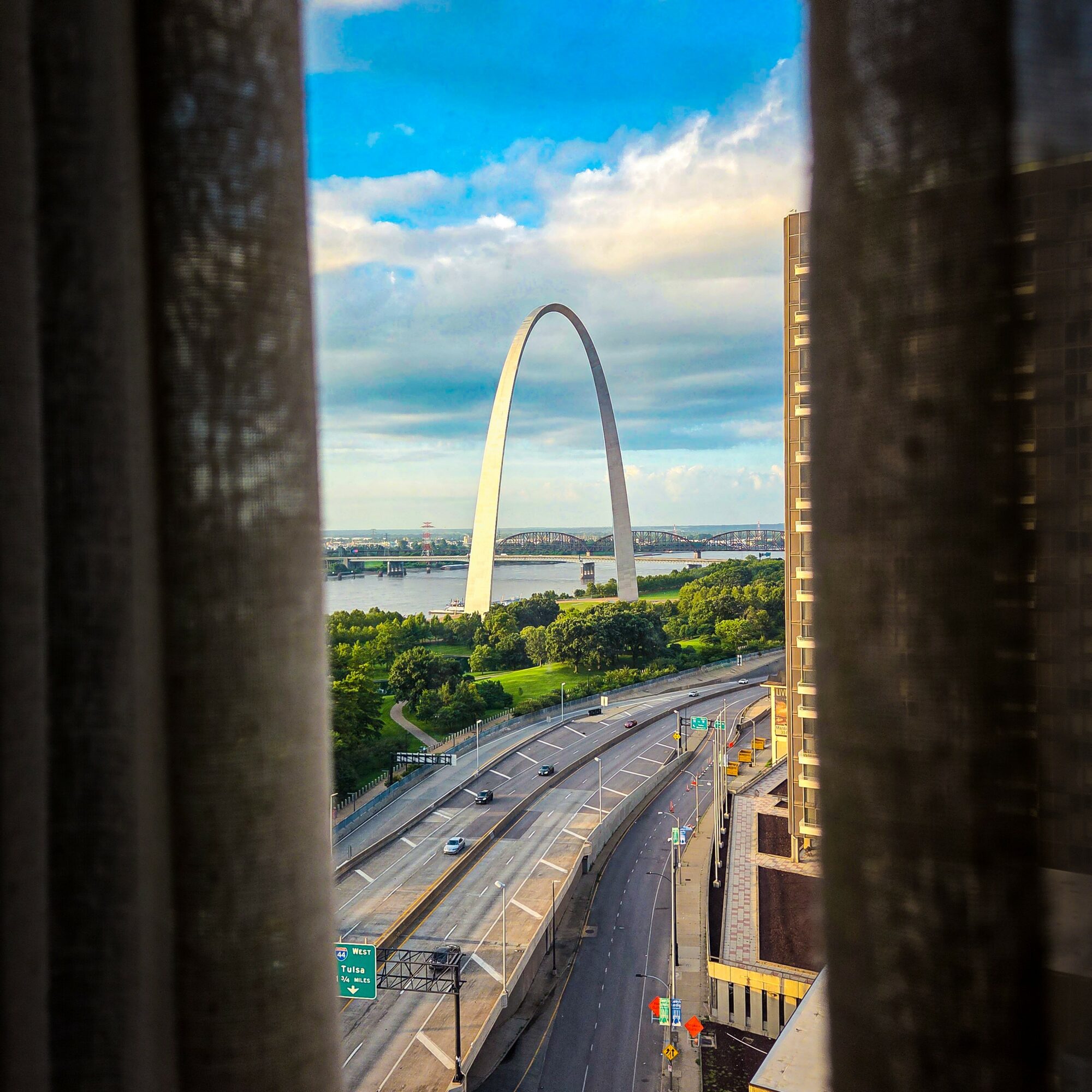 View of the Gateway Arch through a window with curtains, with a highway and cityscape in the foreground.