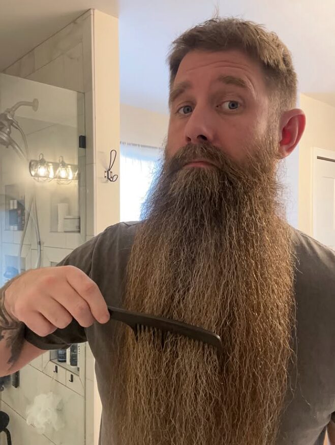 Man with a long beard holding a beard comb, looking at the camera indoors.