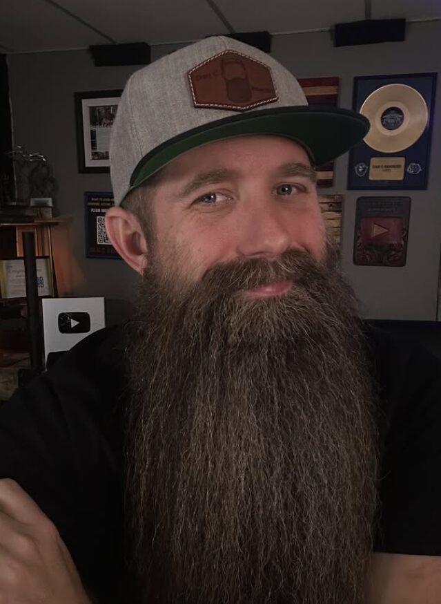 Close-up of a man with a long beard wearing a baseball cap, smiling slightly, in a room with framed awards and pictures