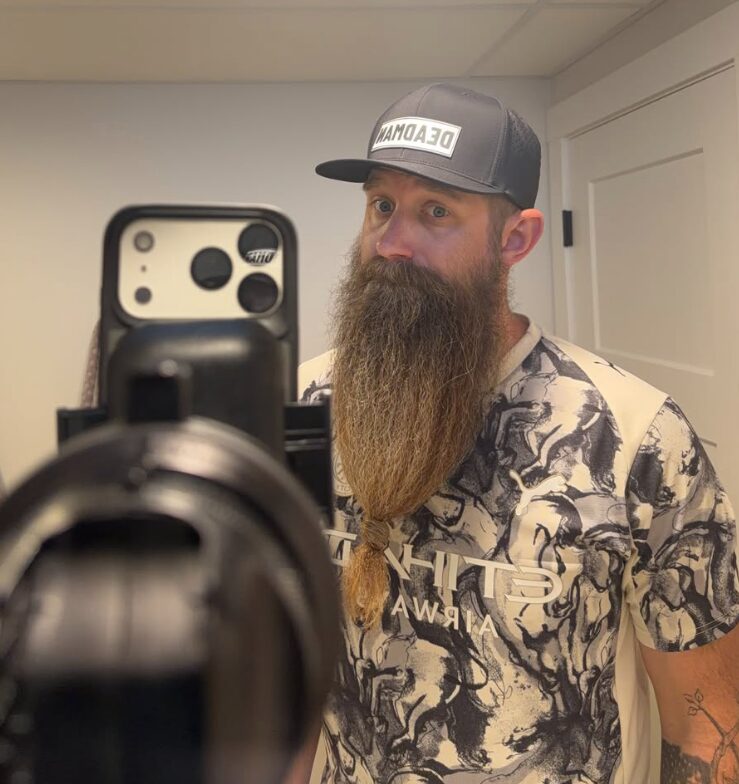 Man with a long beard wearing a cap and patterned shirt, taking a mirror selfie in a room with white walls.