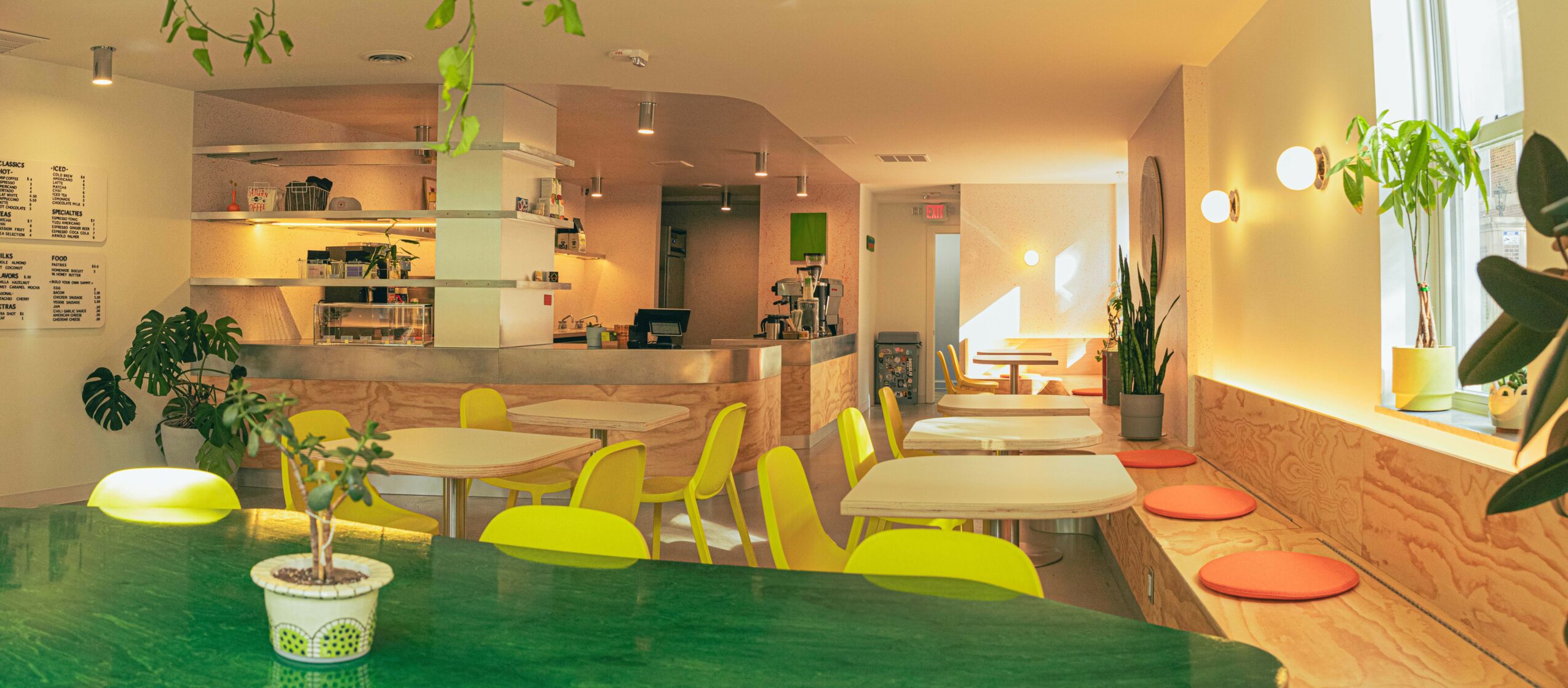 Bright cafe interior with green chairs, wooden tables, potted plants, and large windows letting in natural light.