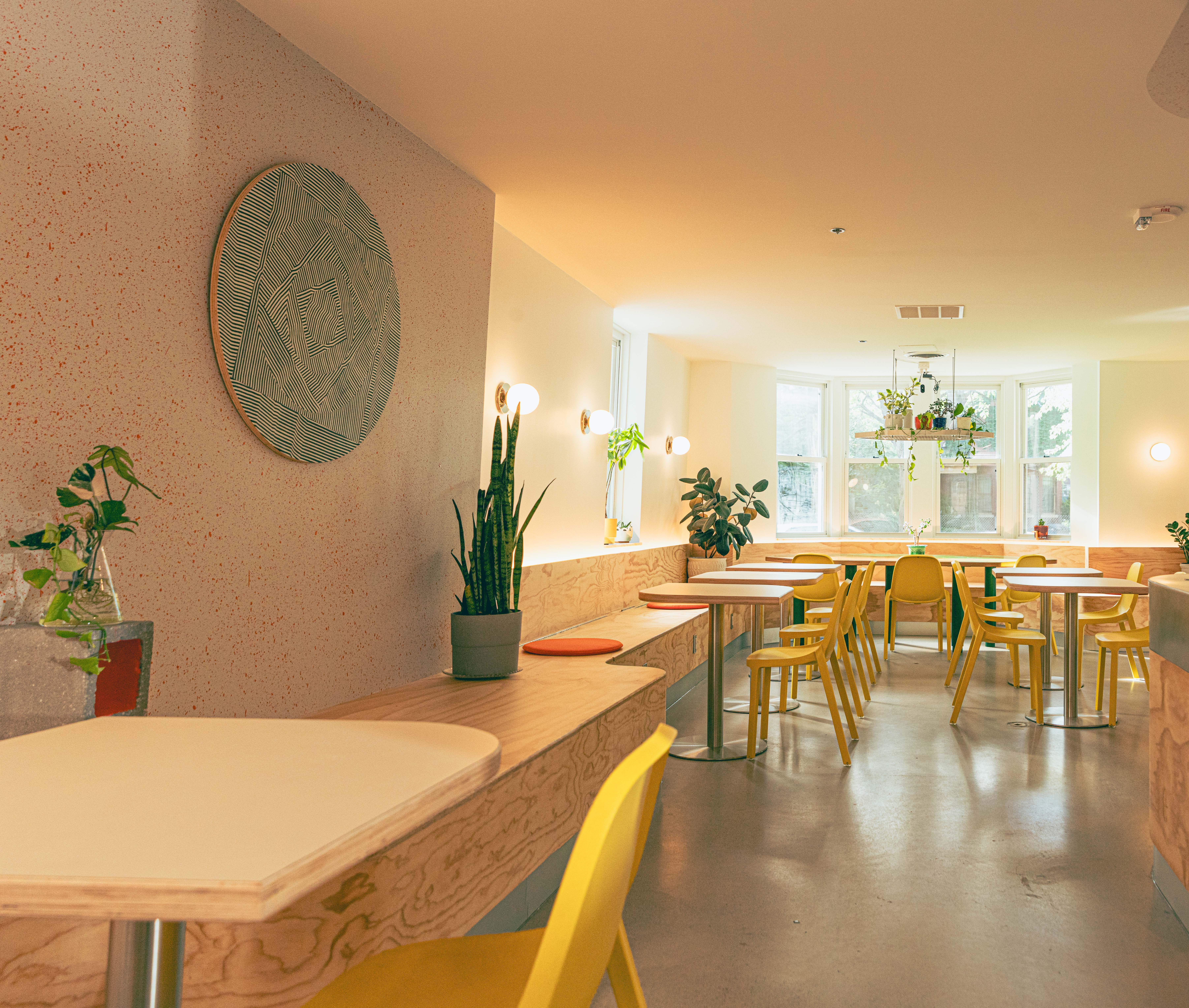 Bright, airy room with yellow chairs, wooden tables, and potted plants near large windows, decorated with a round wall art piece.