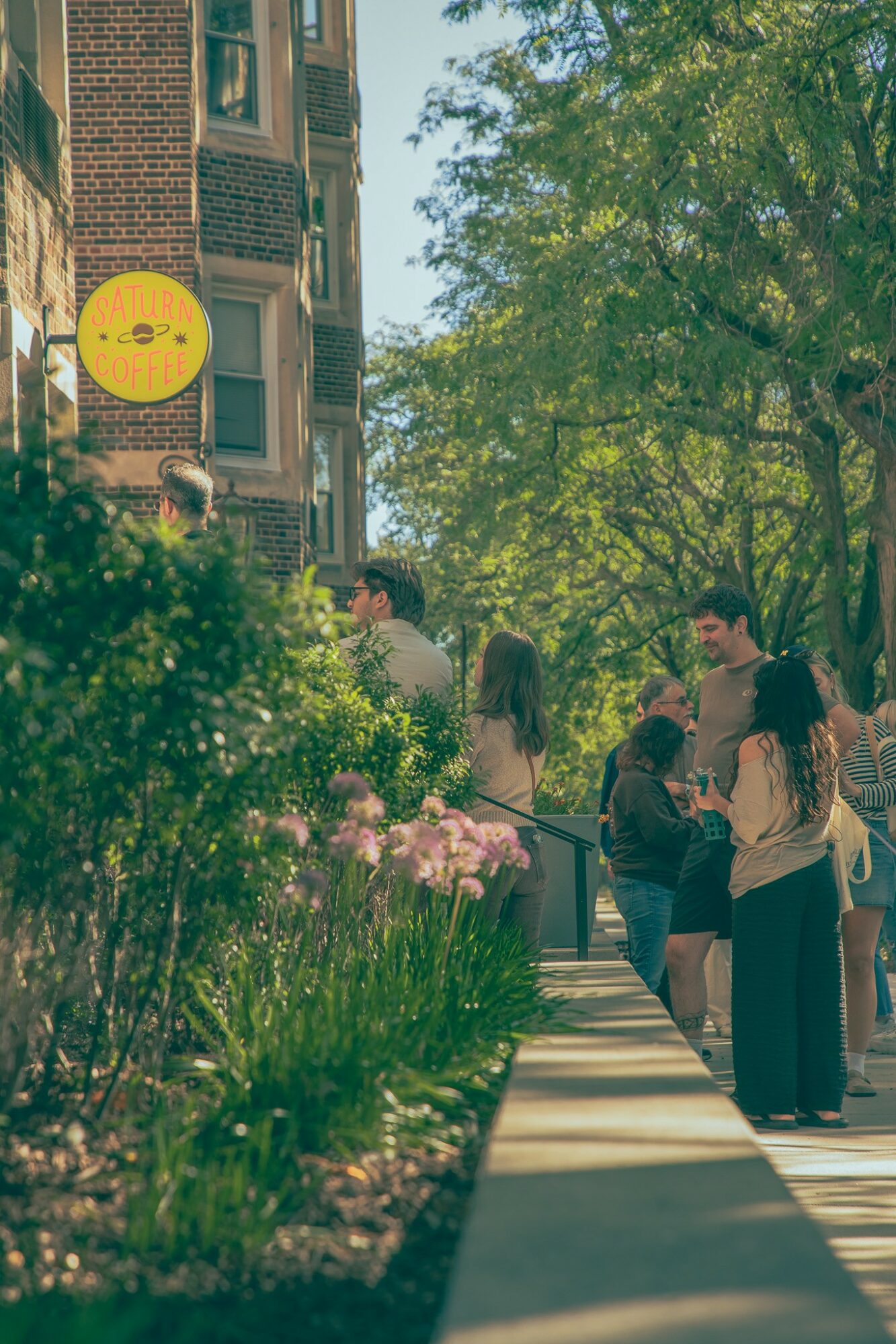 People walking on a sidewalk lined with greenery and pink flowers, with trees and a building in the background.