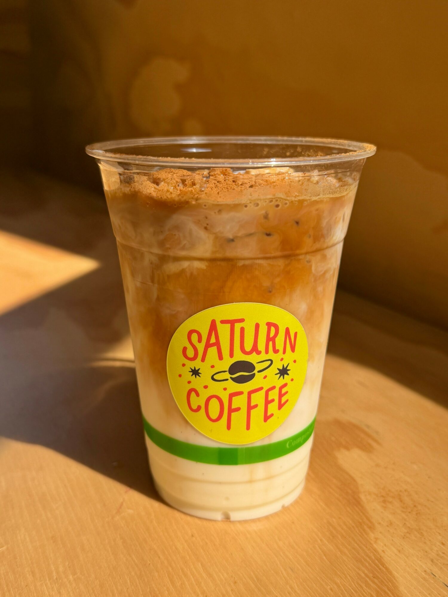 A plastic cup with a yellow and red Saturn Coffee logo, containing a layered iced coffee drink with foam on top.