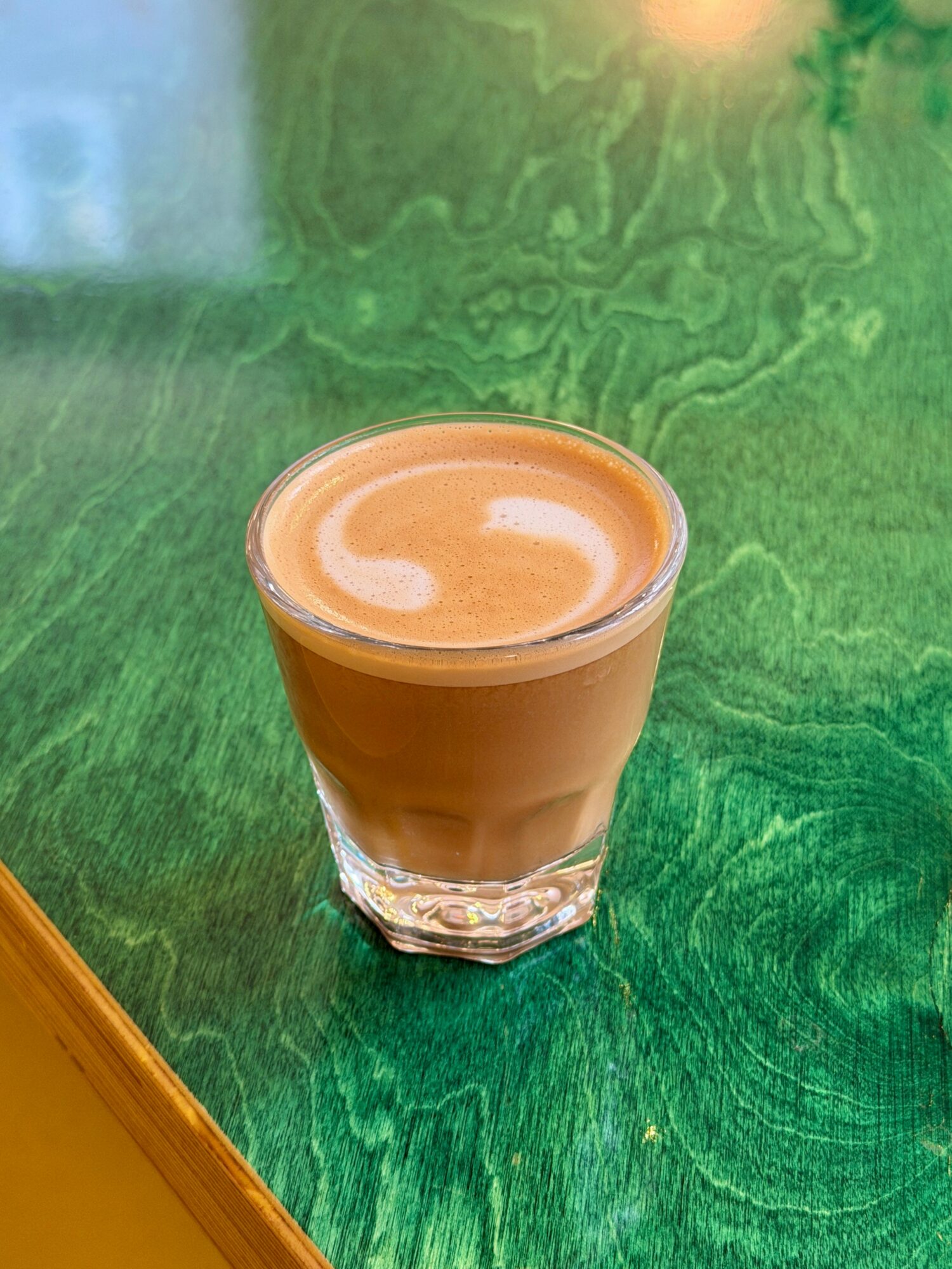 A glass of coffee with foam on top sits on a green textured surface.