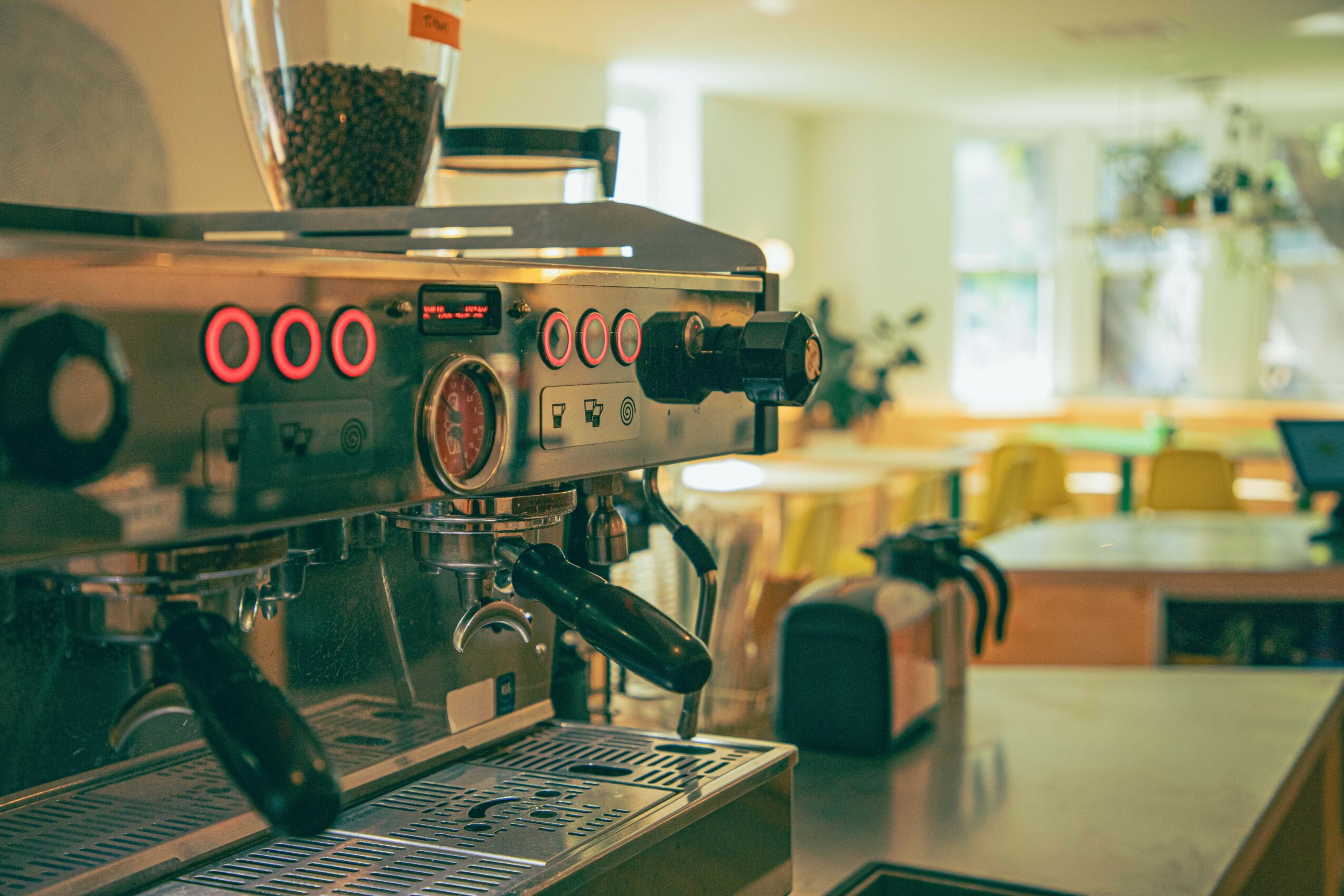 Coffee machine on a counter in a bright cafe with tables and chairs in the background.