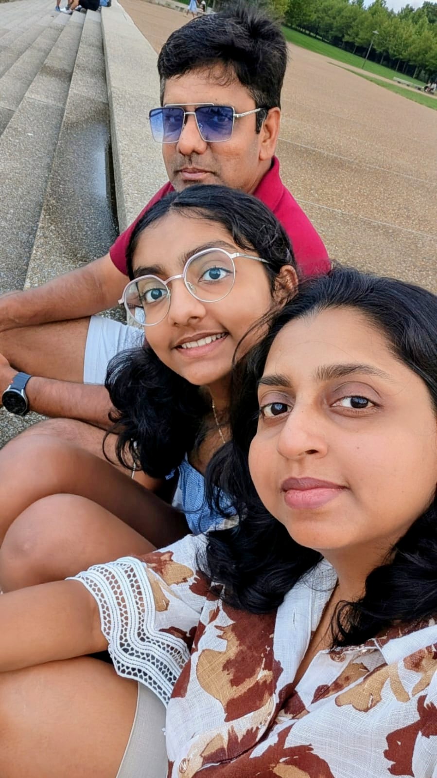 Three people sitting outdoors, smiling, with a park background, wearing glasses and casual clothing.