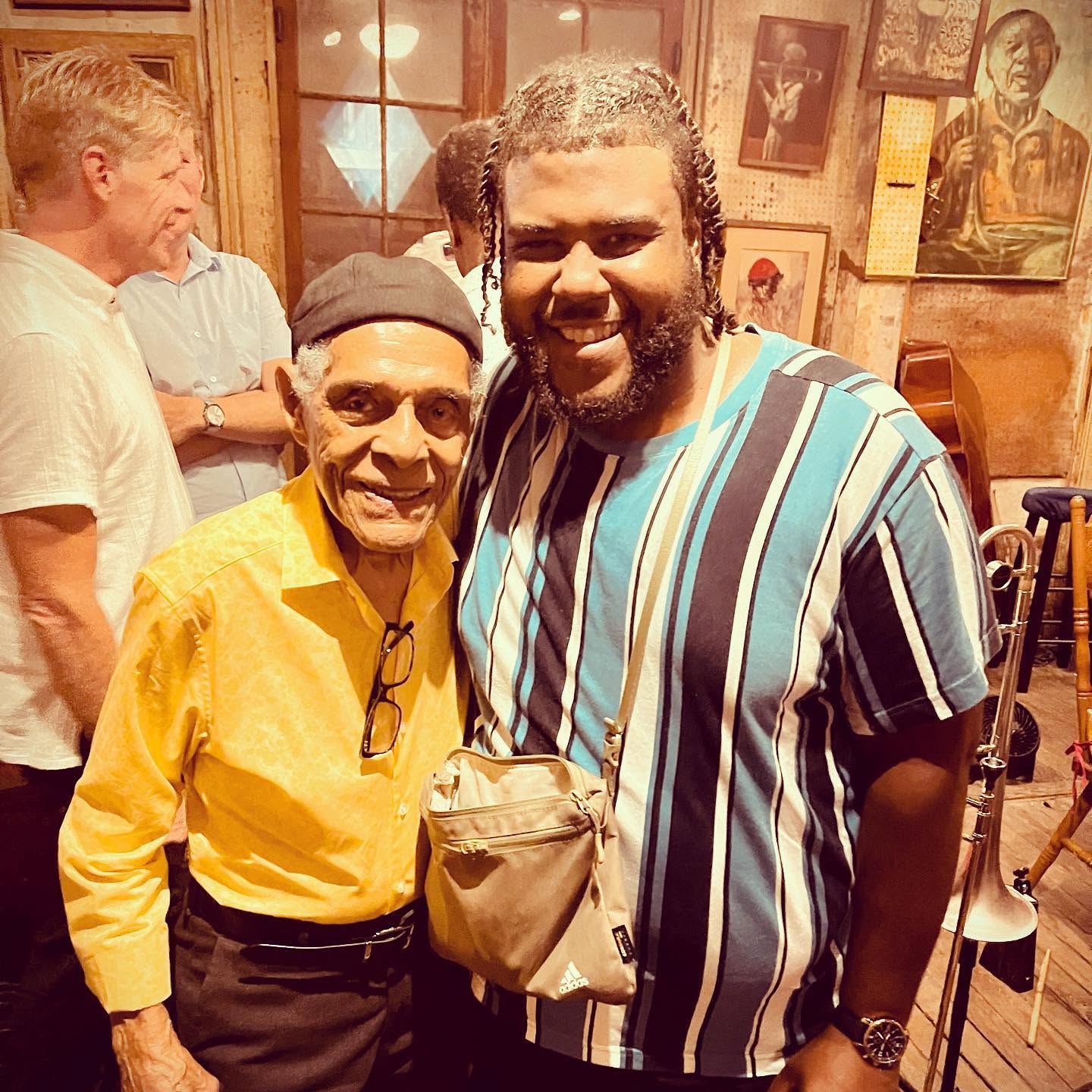 Two men smiling indoors, one wearing a yellow shirt and black cap, the other in a striped shirt, with framed pictures on the wall behind them.