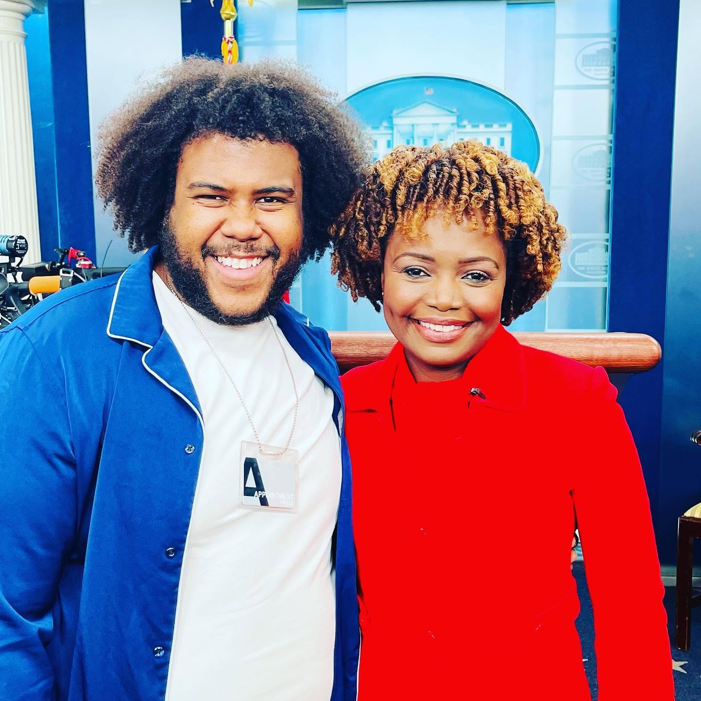 Two smiling people standing close together, one in a blue jacket and the other in a red outfit, in a studio setting.