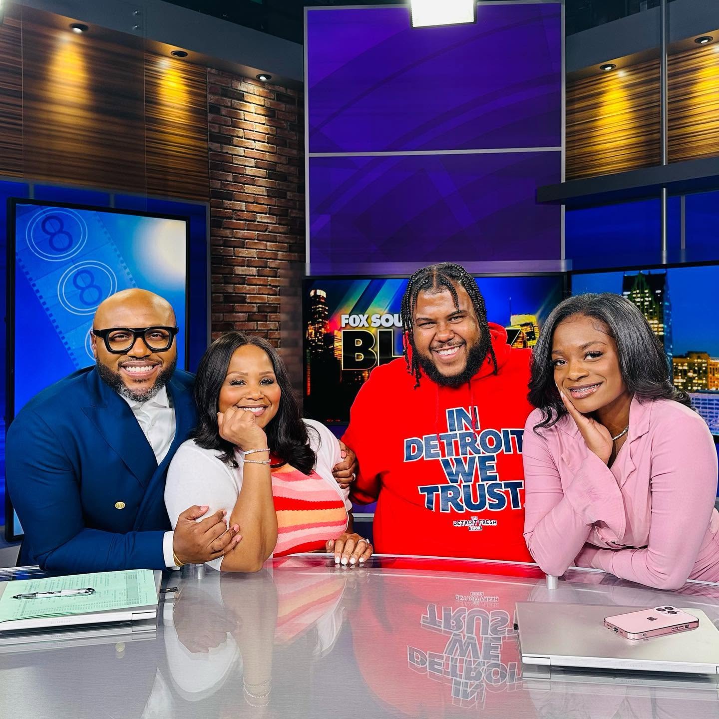 Four people sitting at a news desk in a studio, smiling, with screens and a brick wall background.