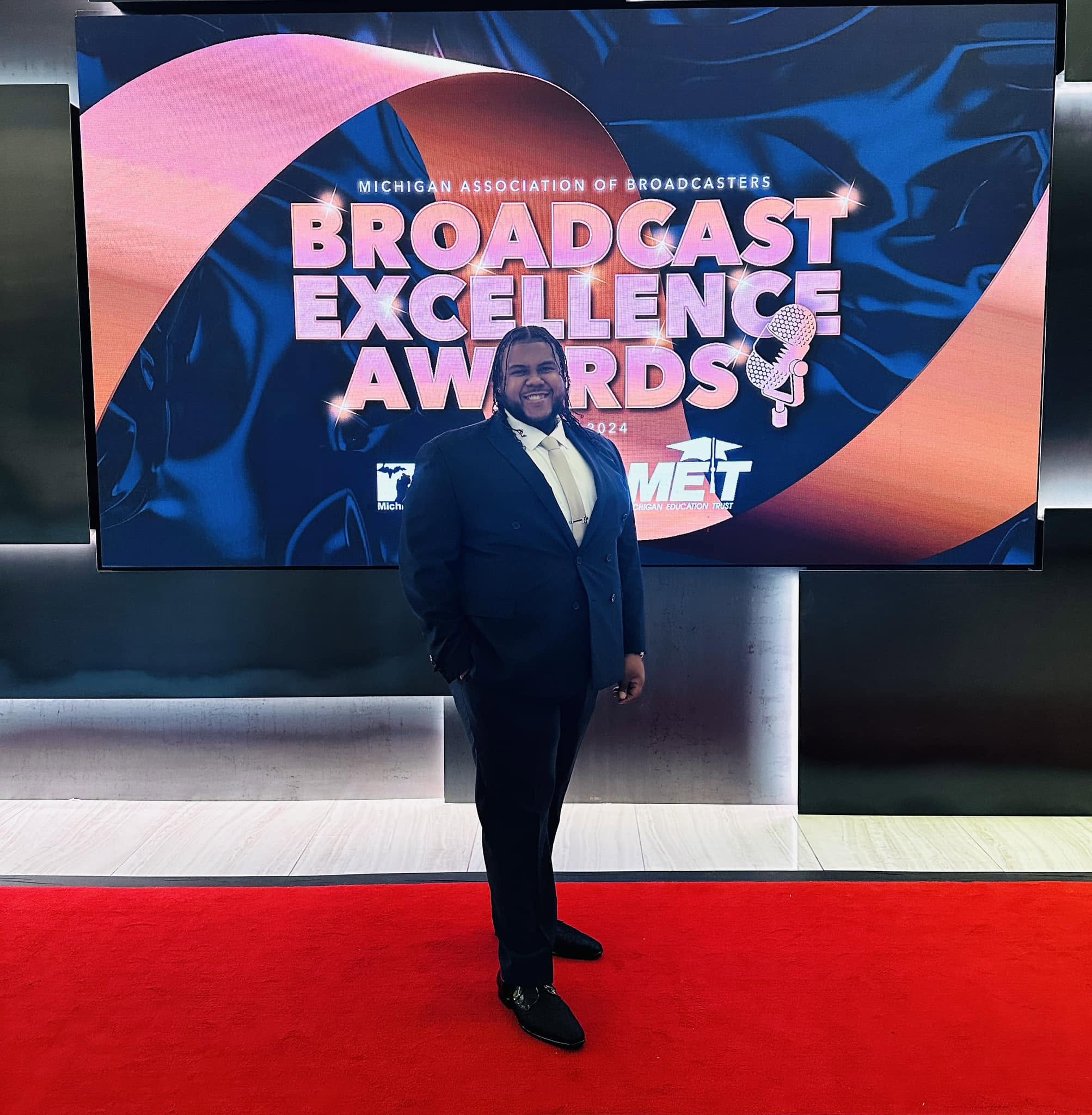 Man in suit standing on red carpet in front of a large screen with event text, smiling, indoors.
