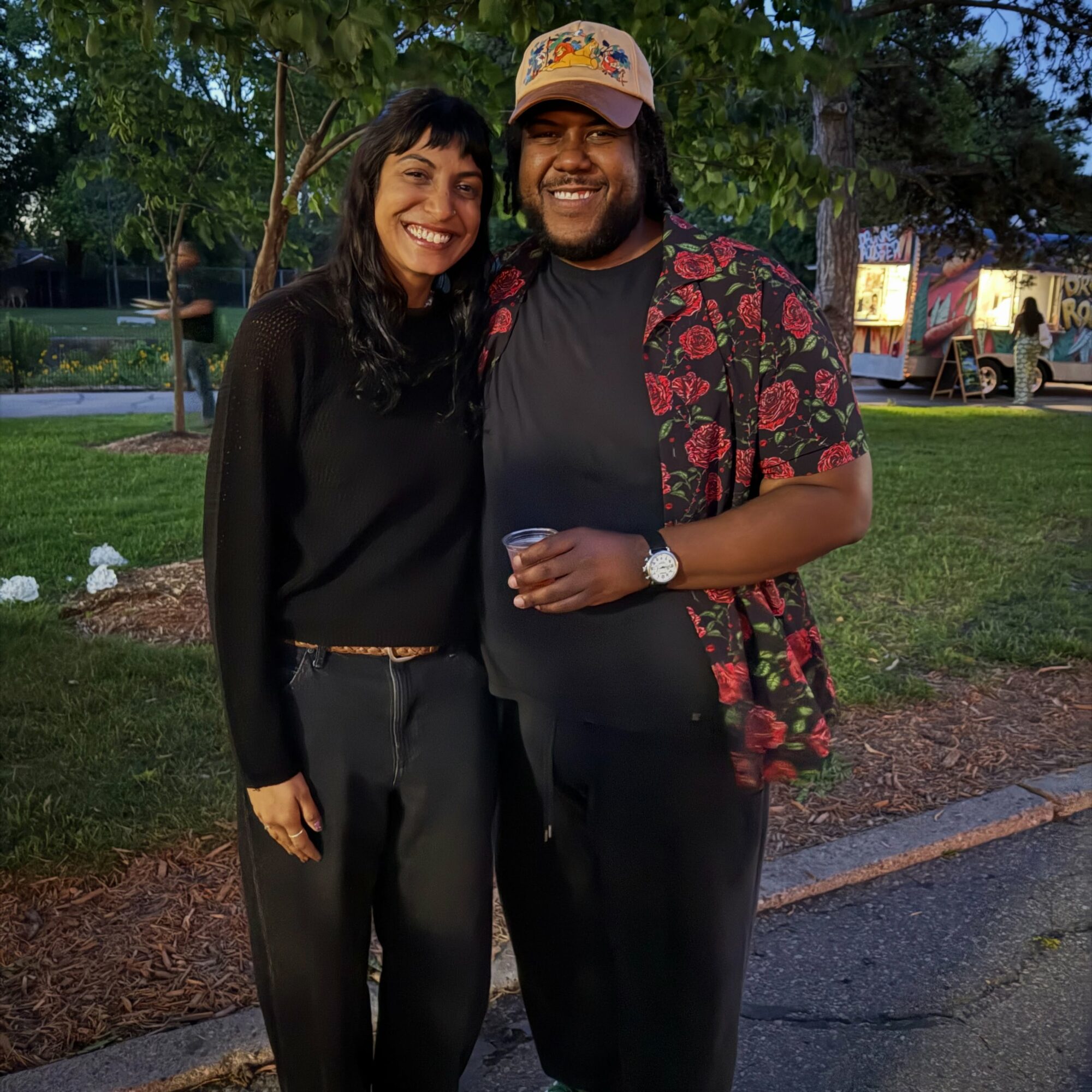 Two people smiling outdoors at night, standing on grass with trees and a lit-up area behind them.