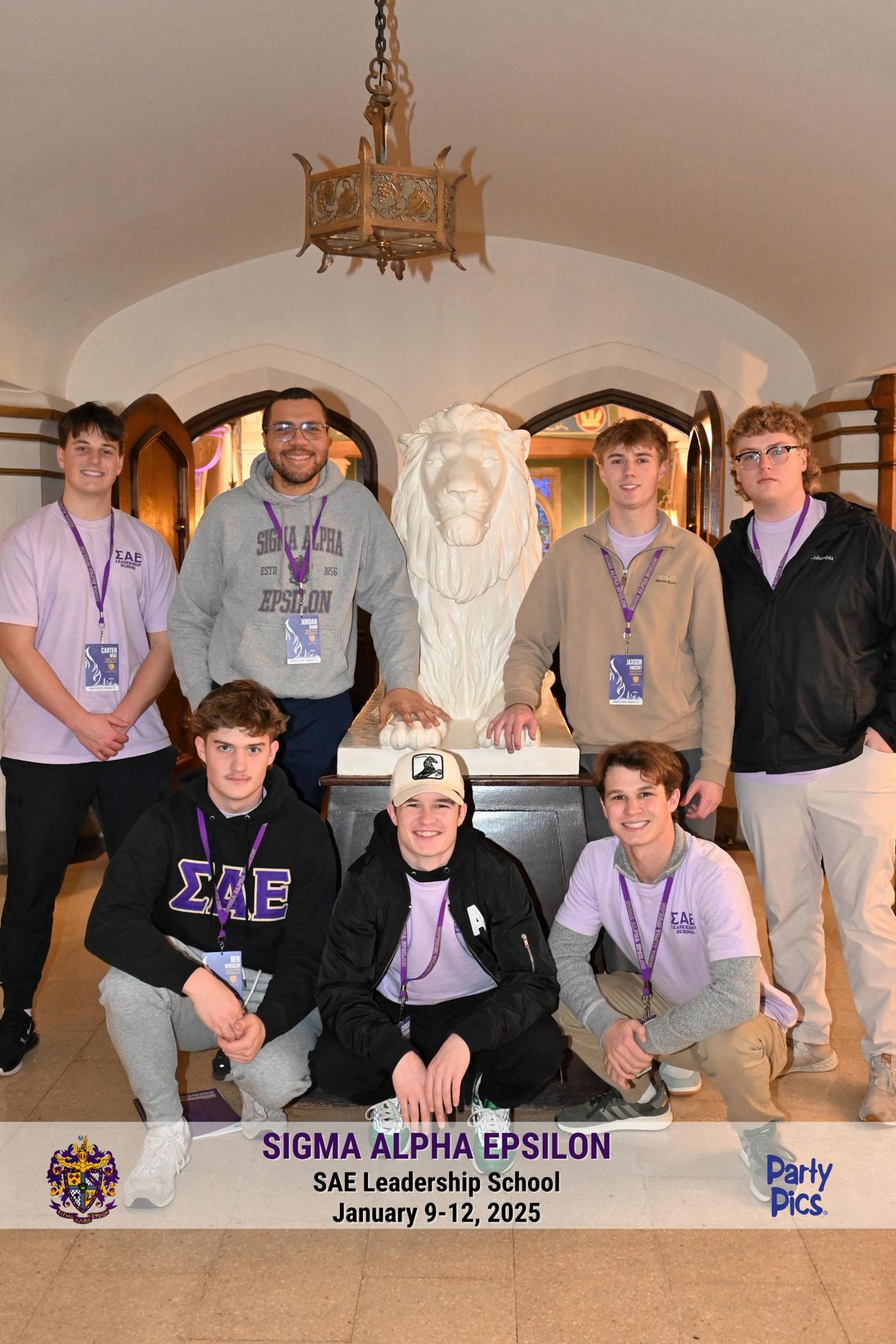 Group of seven people, five standing and two sitting, in a room with a lion sculpture and a chandelier, holding a Sigma Alpha Epsilon banner.