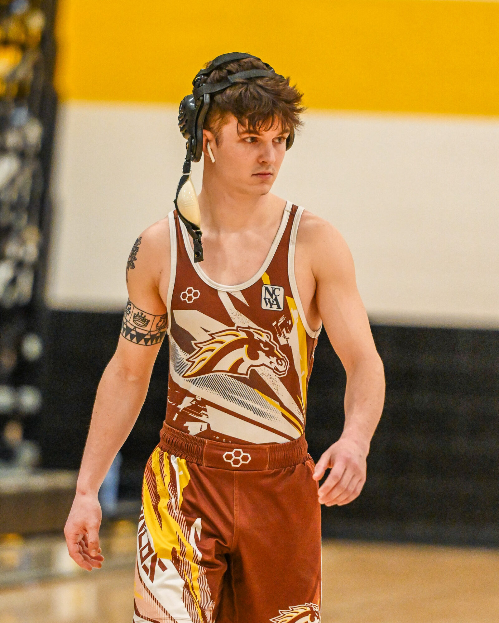 Female wrestler in singlet and headgear standing on wrestling mat, with a yellow and white wall in background.