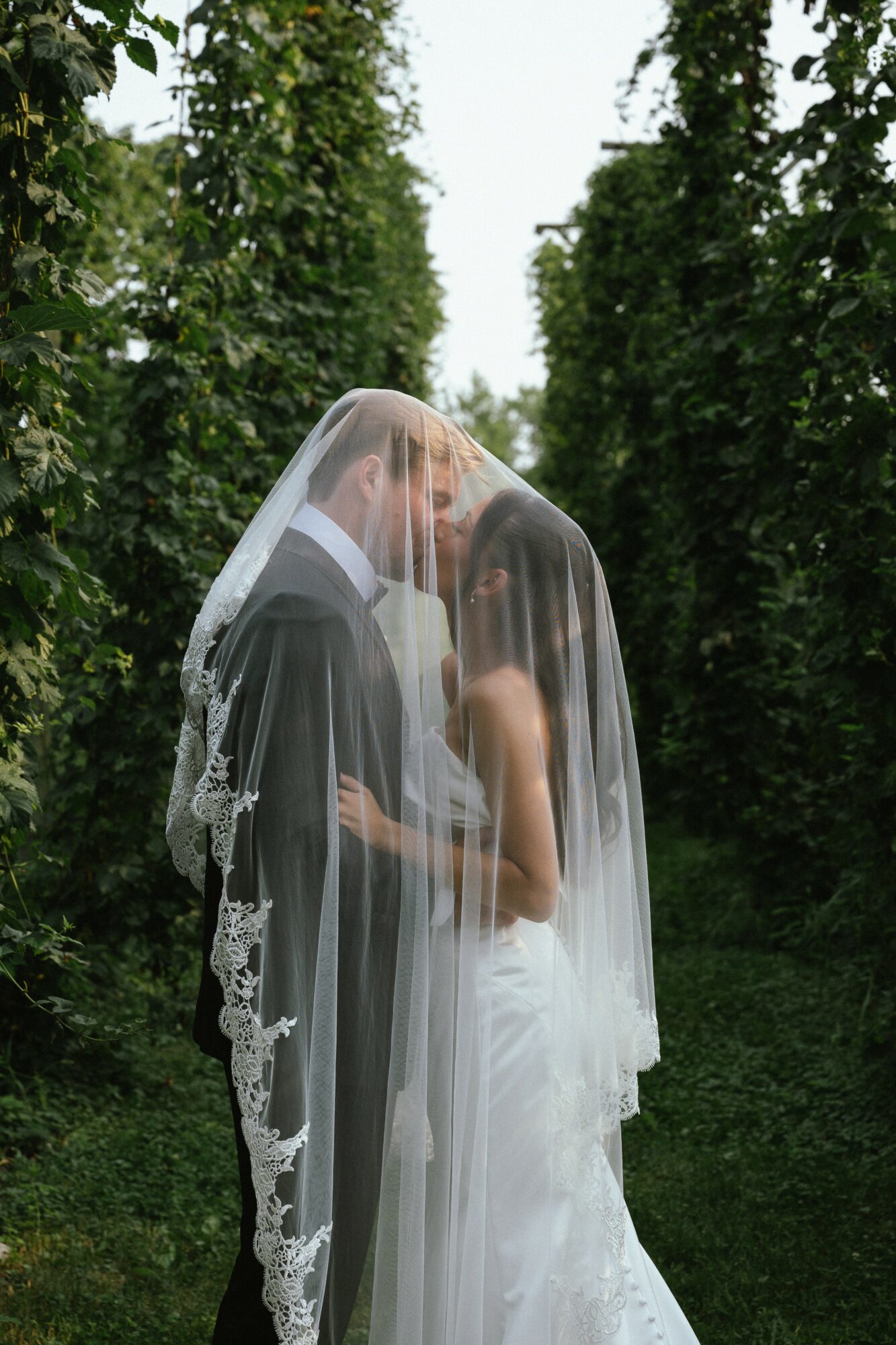 Bride and groom standing close, with the bride's veil covering both, in a green outdoor setting.