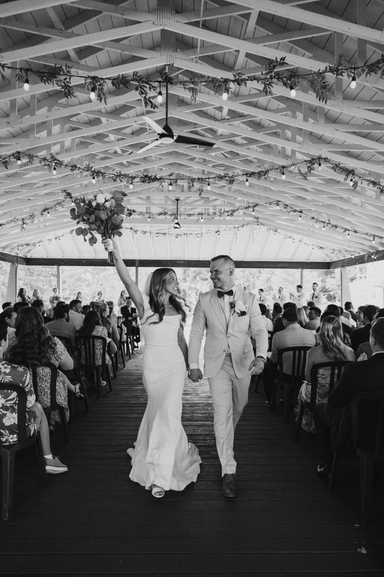 Couple holding hands walking through decorated event space with seated guests and floral arrangements, ceiling with draped fabric and hanging greenery.