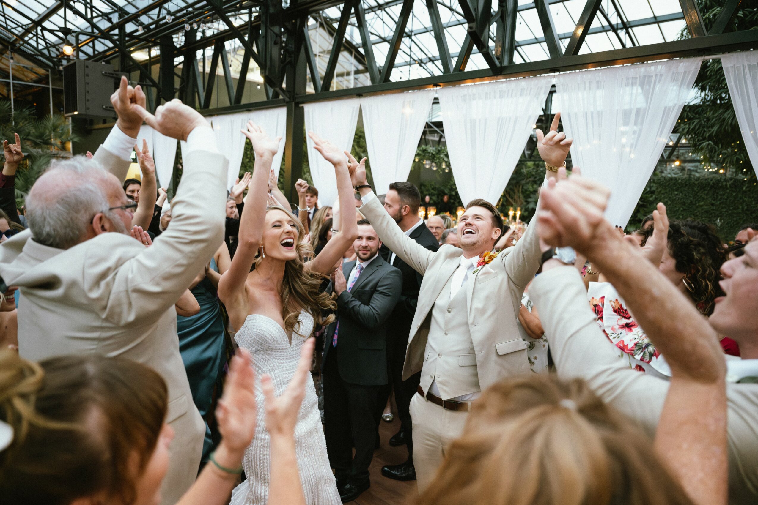 Group of people celebrating indoors, smiling, raising hands, and dancing at a wedding reception.
