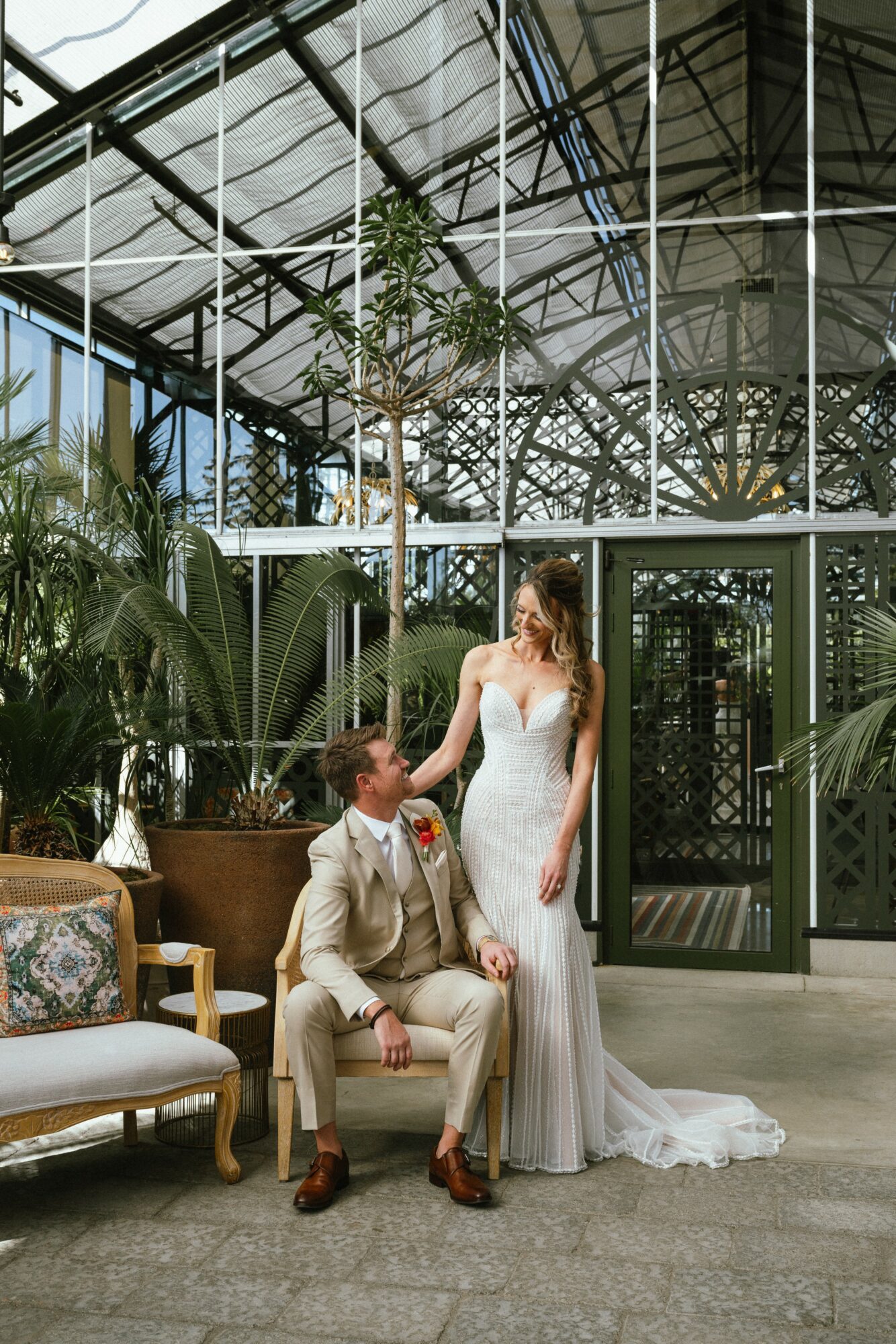 A bride in a white wedding dress and a groom in a beige suit are in a greenhouse with large plants and glass walls.