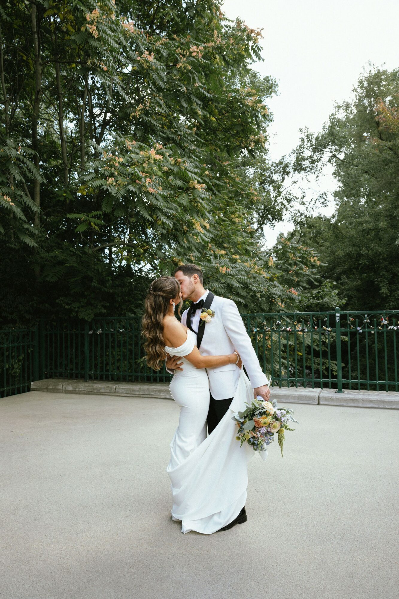 A couple in wedding attire standing outdoors, embracing and kissing, with trees and a fence in the background.