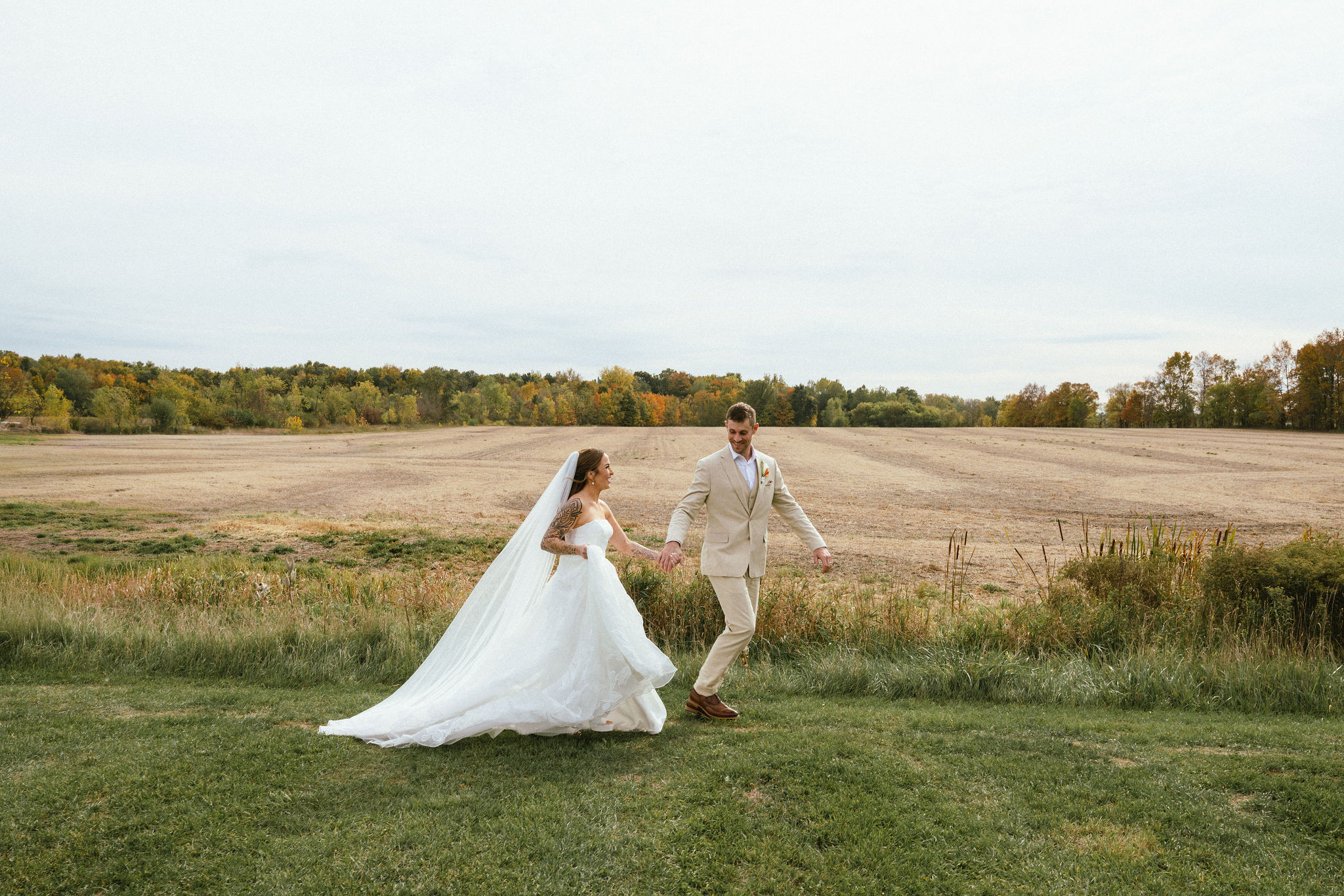 A bride and groom holding hands walk across a grassy field with a distant landscape and cloudy sky.