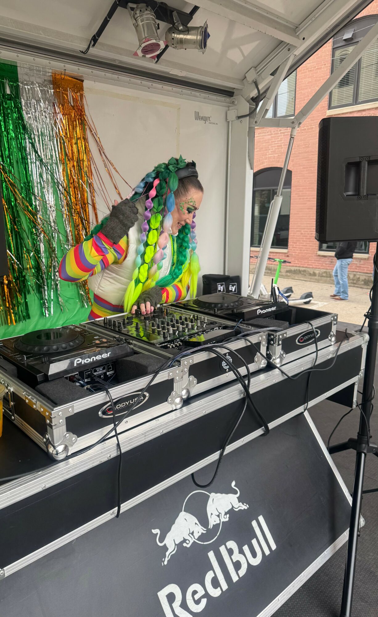 Person with colorful braided hair and rainbow striped sleeves DJing at an outdoor event, with a Red Bull logo on the table.