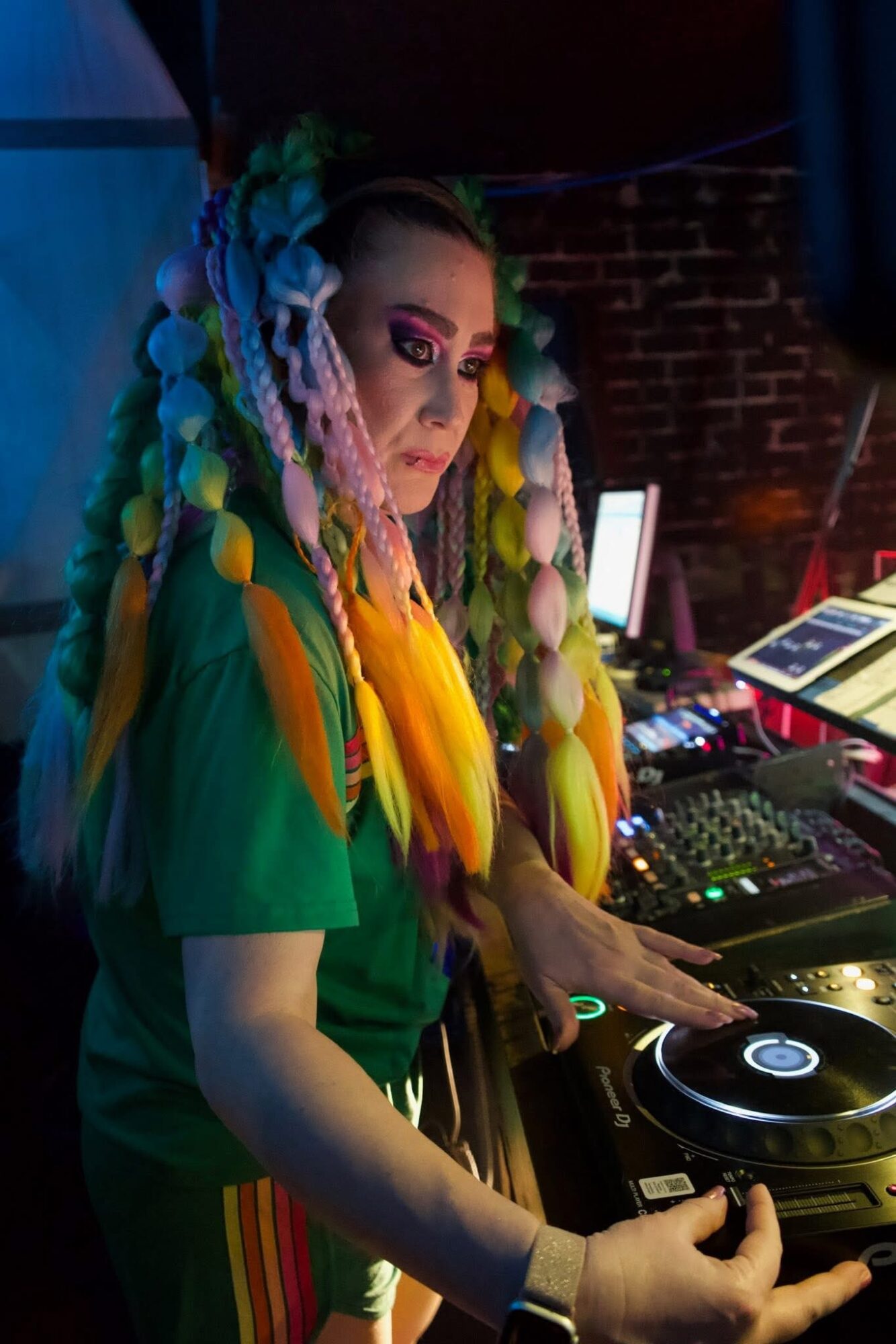 Female DJ with colorful braided hair operating DJ equipment in a dimly lit venue.