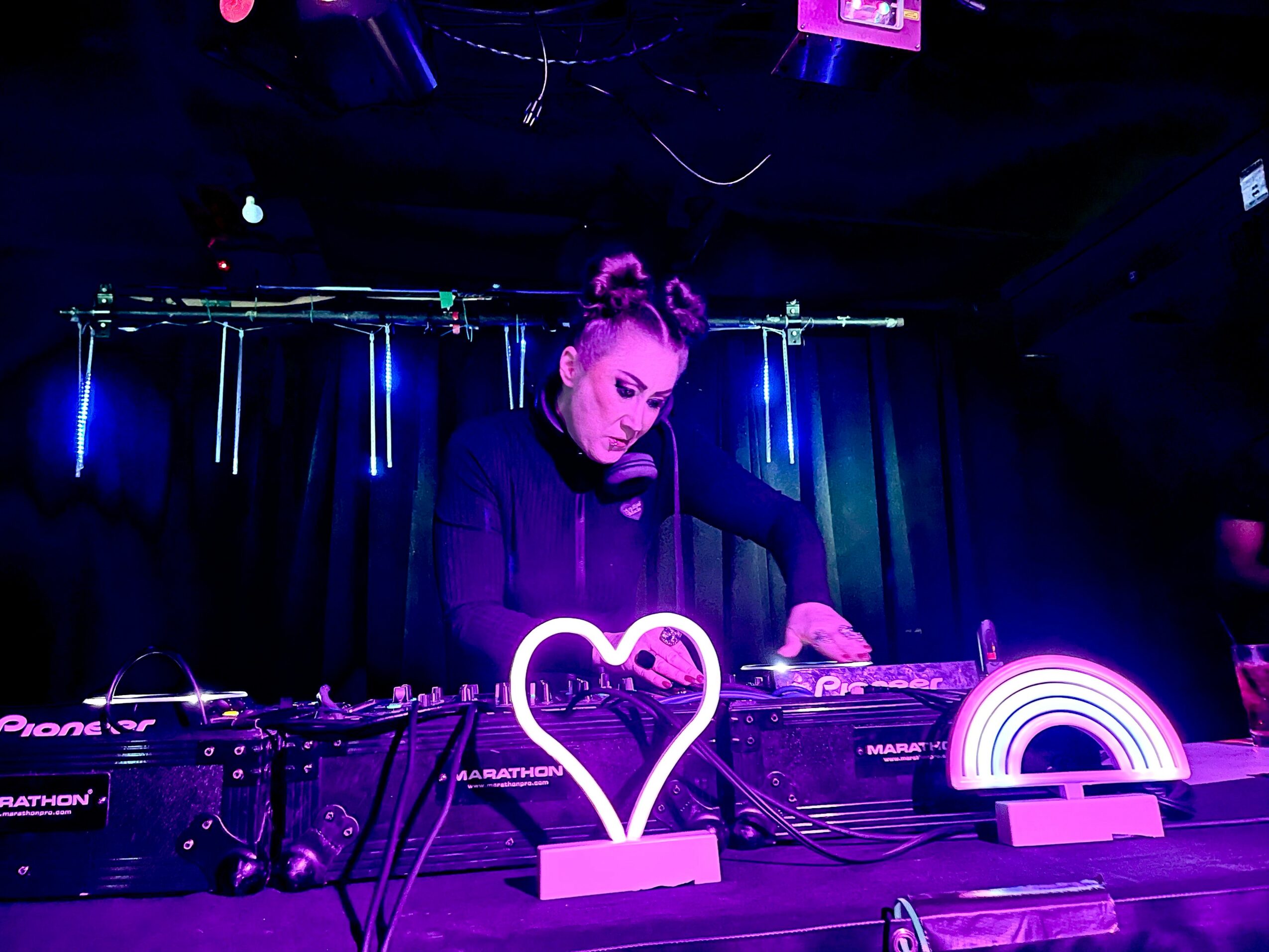 Female DJ with hair in buns performs at a neon-lit event, surrounded by DJ equipment and colorful lights.