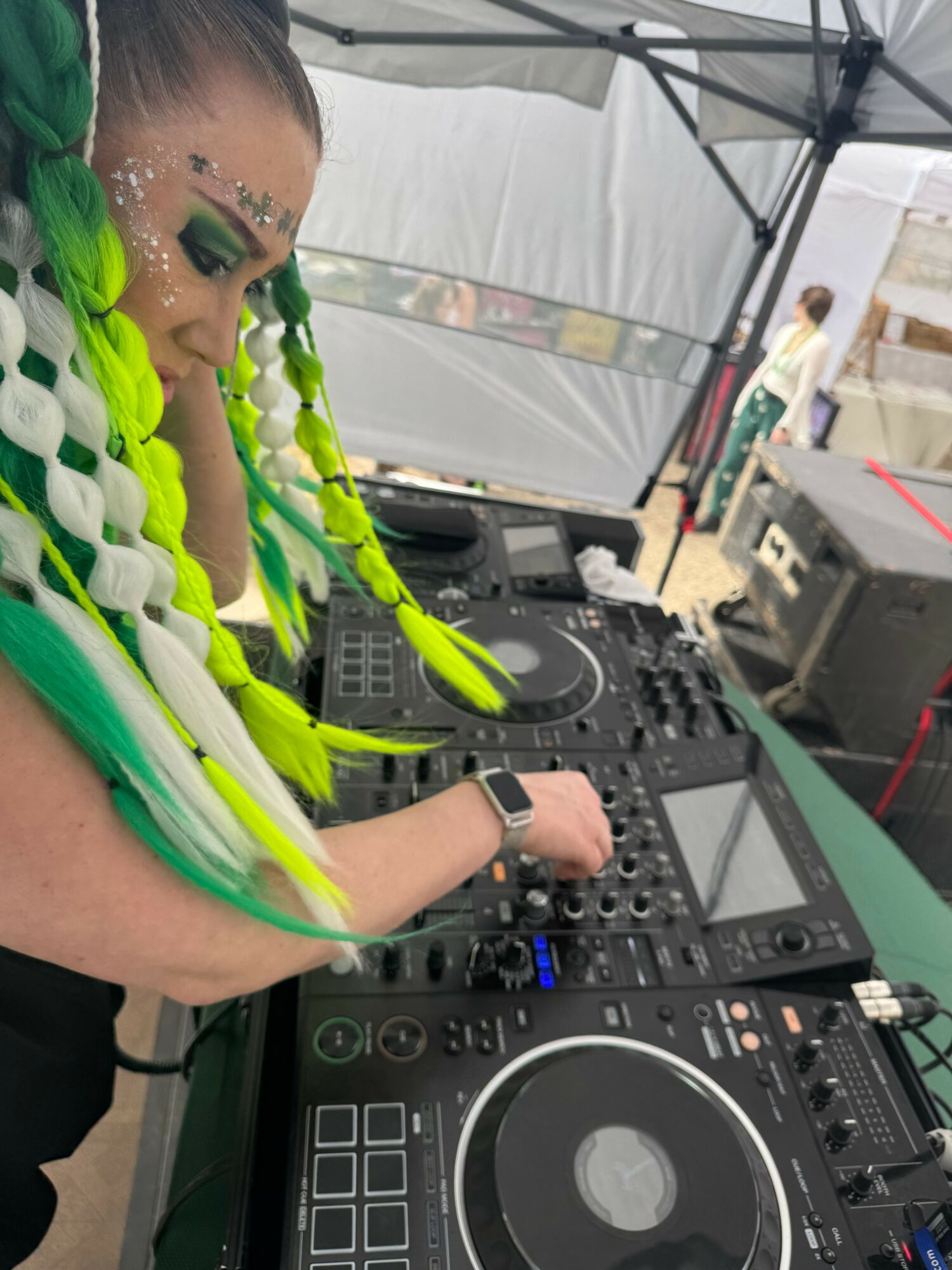 Person with colorful braided hair using DJ equipment outdoors, with a tent and equipment in the background.