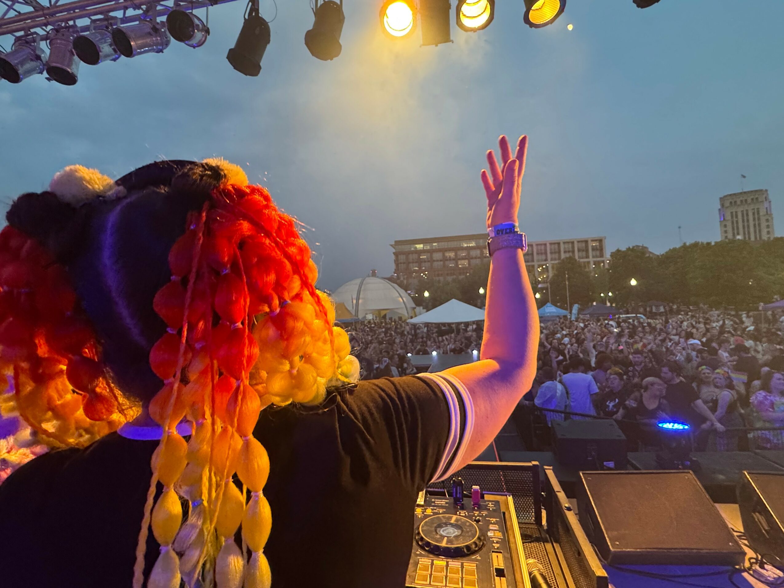 Person with colorful hair raising hand on stage at outdoor concert, audience and cityscape in background.