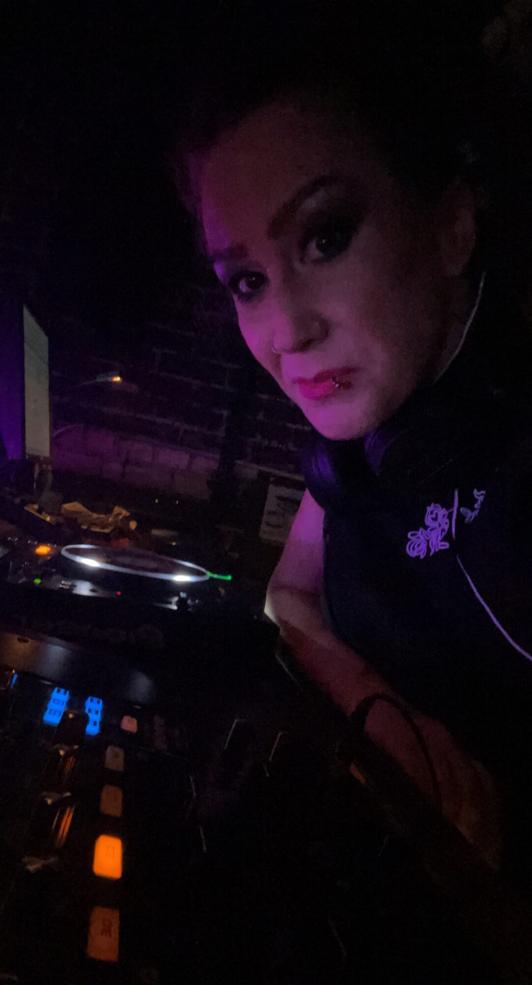 Person with dark hair and makeup, DJ equipment in a dark setting with purple lighting.