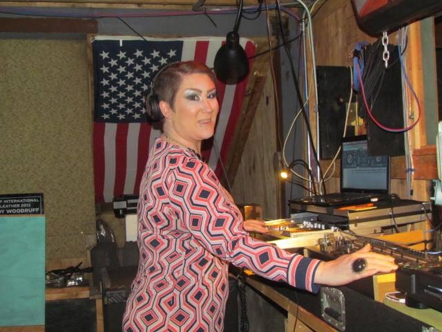 Woman with makeup and styled hair operating DJ equipment in a room with an American flag in the background.