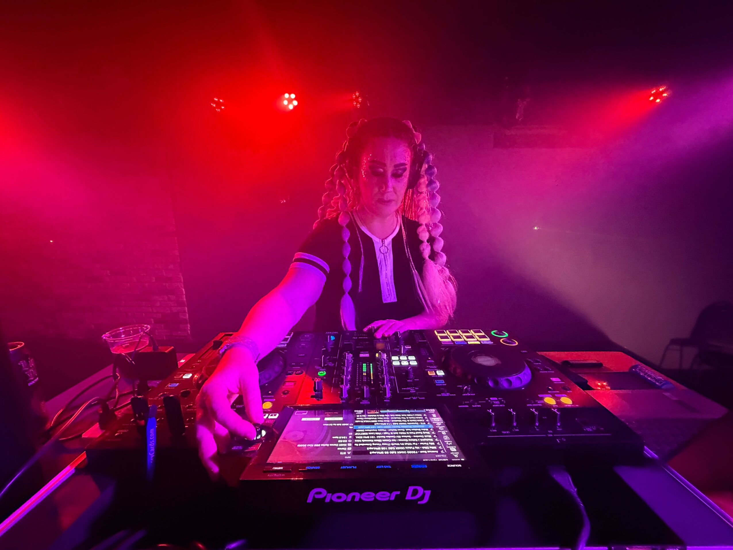 Female DJ with curly hair and headphones, operating DJ equipment in a colorful, dimly lit setting.