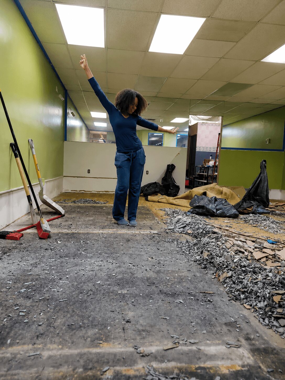 Child with curly hair in a blue shirt and jeans raises arms in a room under renovation, with construction tools and debris.