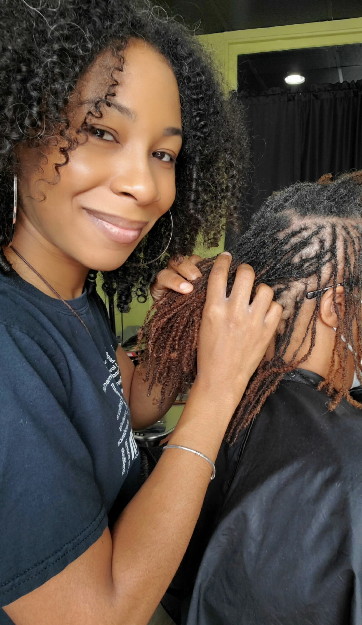 Woman with curly hair smiling, touching another person's dreadlocks, in an indoor setting.