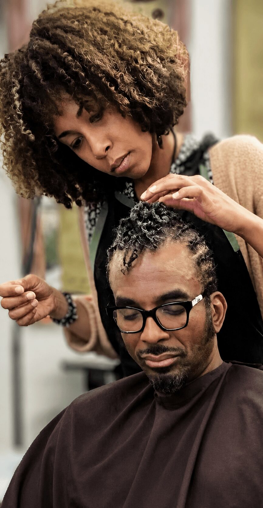 Woman styling a man's hair in a salon, woman has curly hair, man wears glasses, both focused on hair care.
