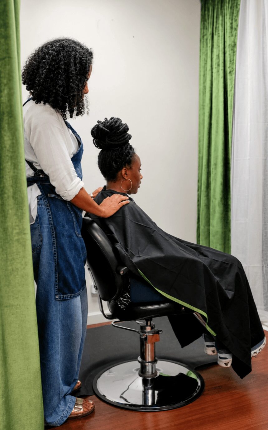 A person with curly hair stands beside a seated woman with an updo hairstyle in a salon, with green curtains in the background.