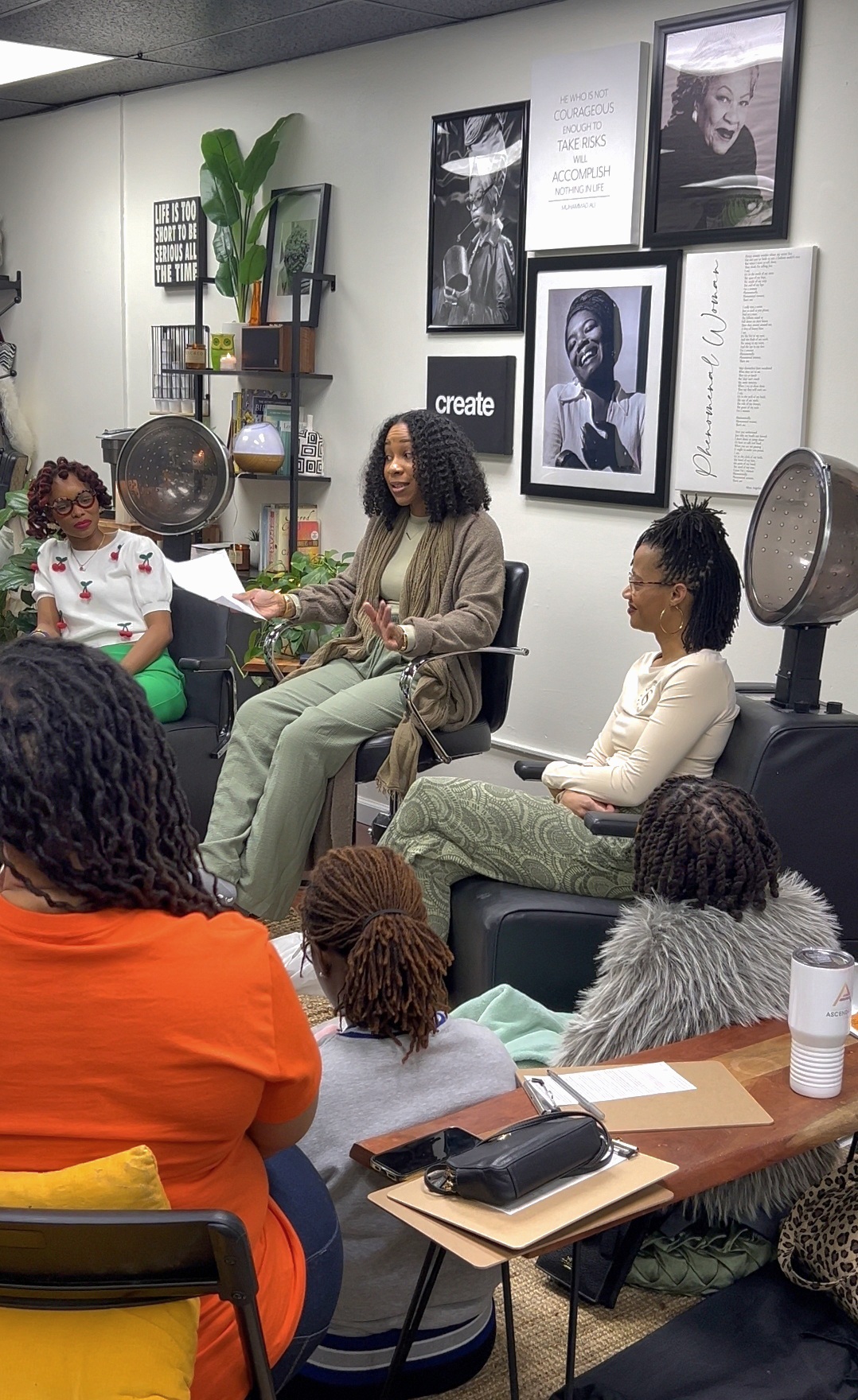 Group of people seated in a salon, with hairdryers and framed pictures on the wall behind them.