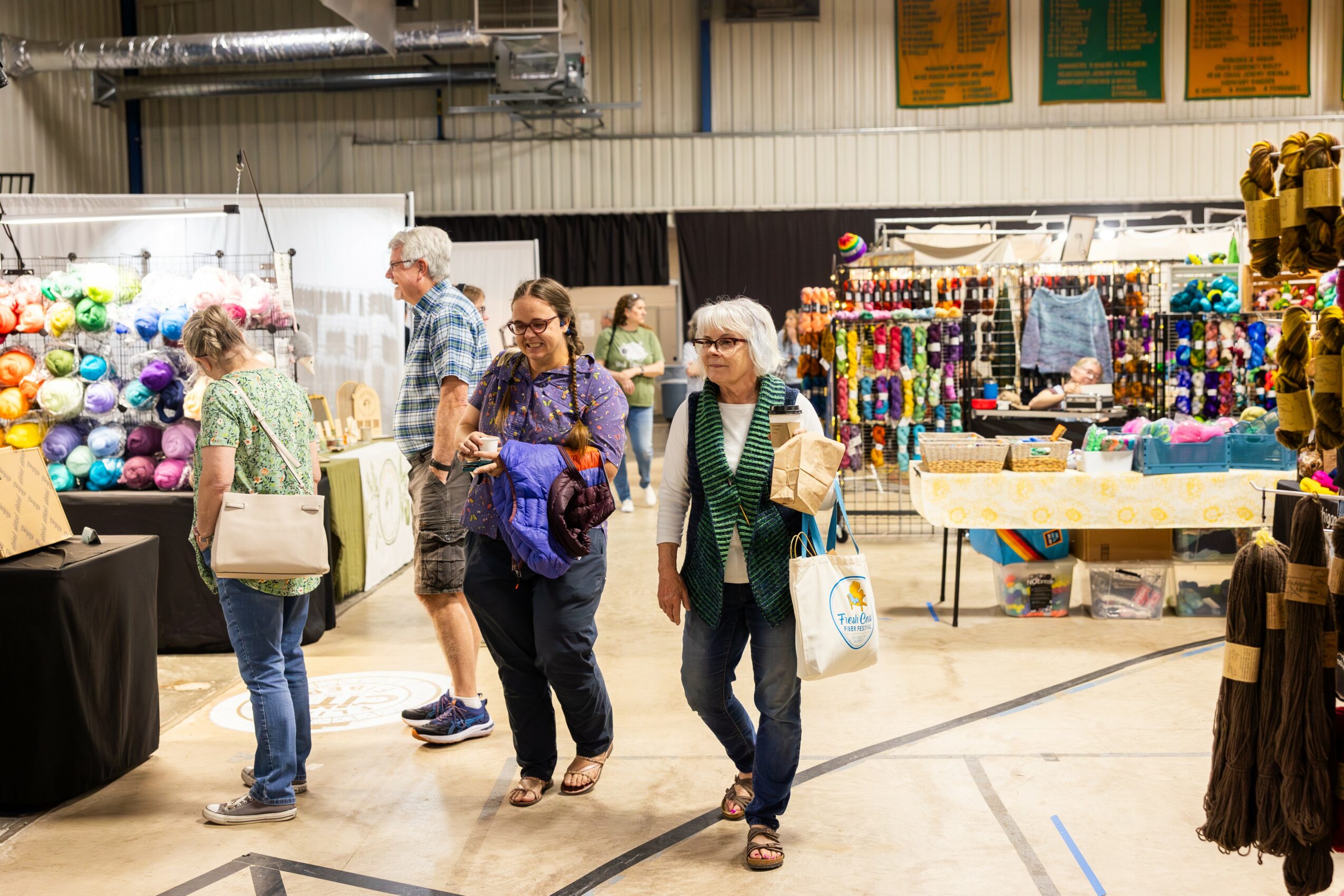 People browsing craft supplies and yarn at an indoor market, with tables and colorful items displayed.
