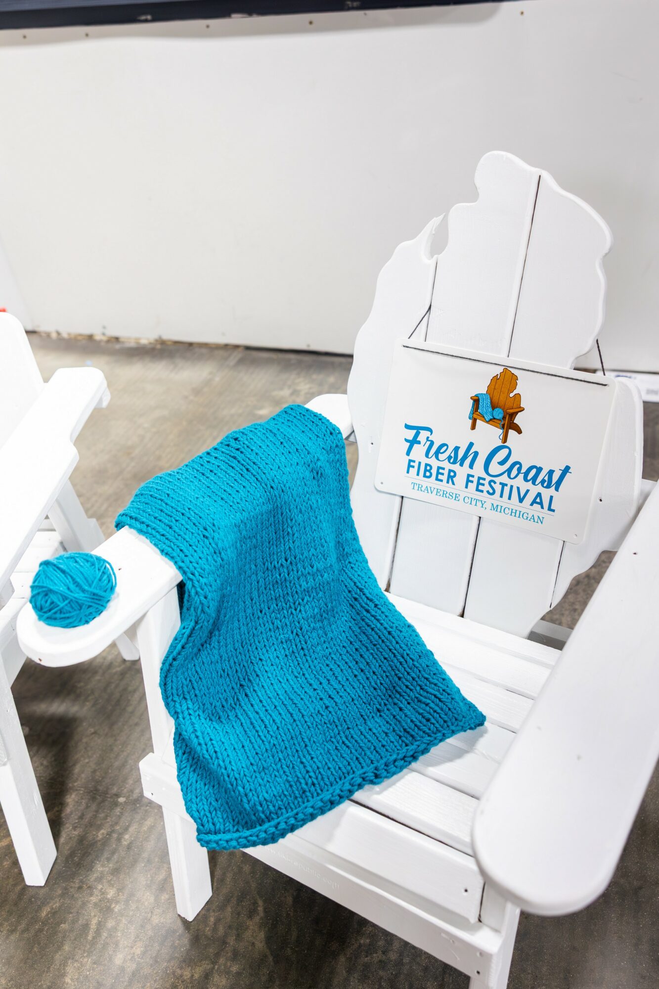 White Adirondack chair with a blue knitted blanket draped over it, advertising the Fresh Coast Fiber Festival, outdoors.