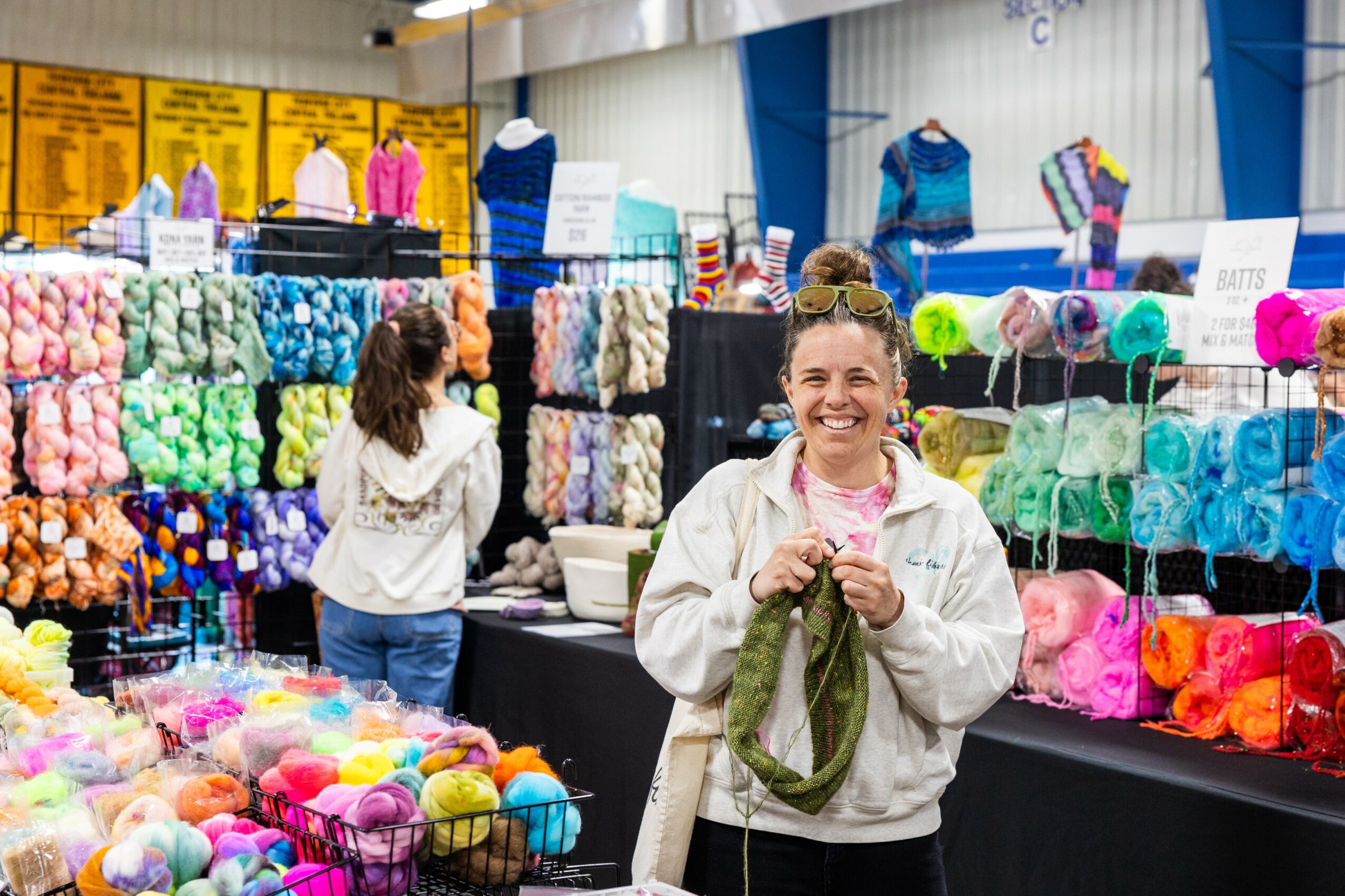 Smiling woman holding yarn at colorful yarn display in a craft fair, with another person browsing in the background.