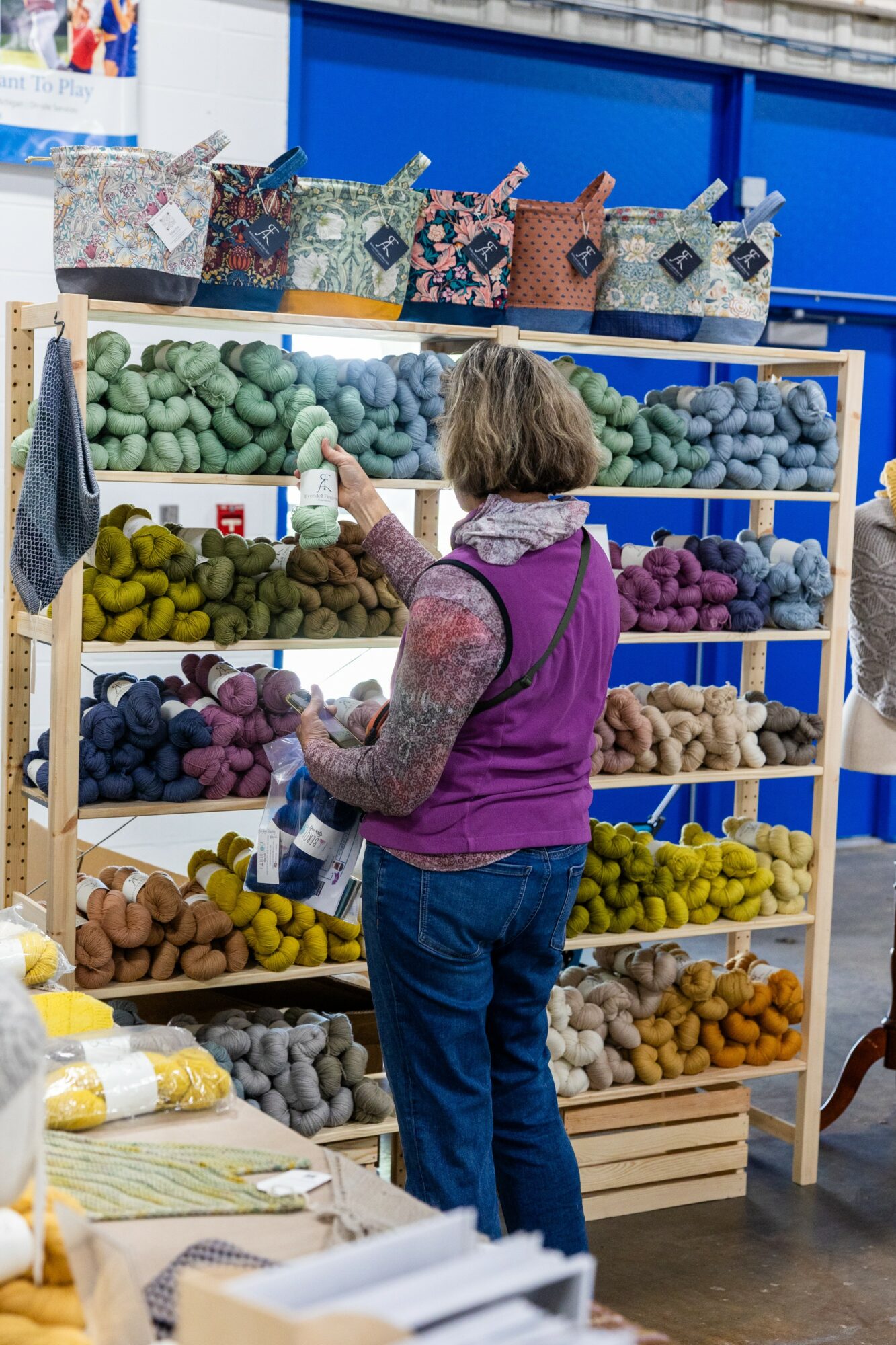Woman shopping for yarn in a store, shelves filled with colorful yarn balls, wearing a purple vest and jeans.